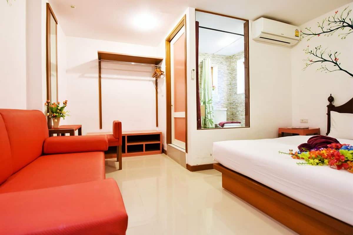 Una habitación de hotel con una cama, un sofá rojo, una mesita con un jarrón de flores, un ventanal con vistas a zonas verdes y un equipo de aire acondicionado.