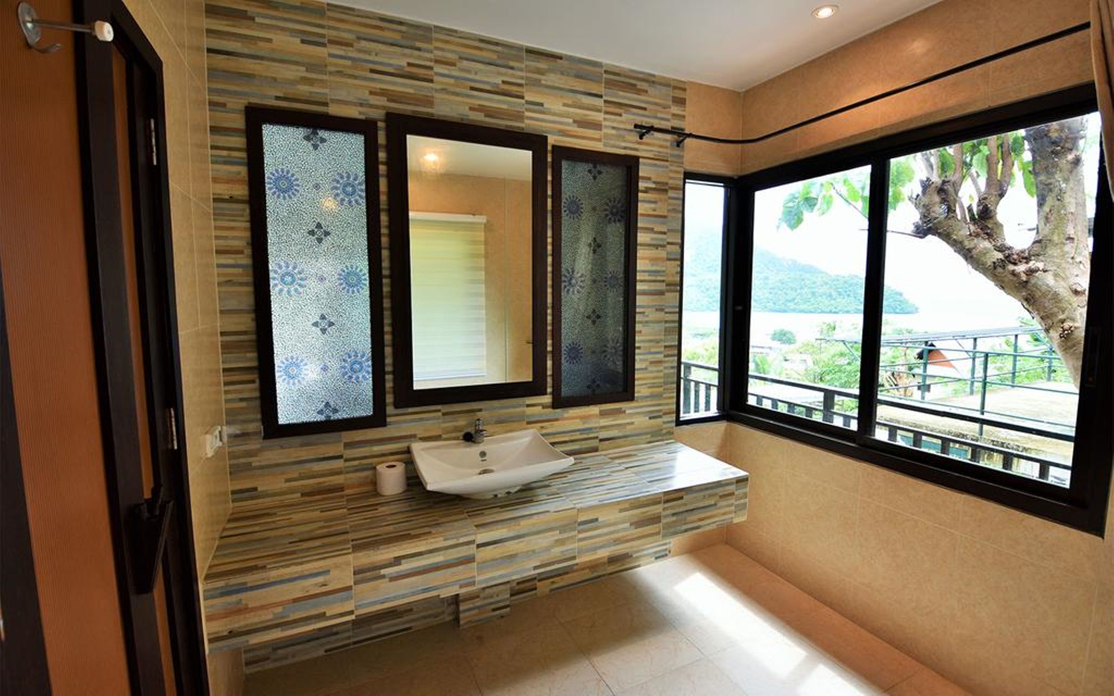 A bathroom with a sink, three mirrors, and a view of a tree and landscape through large windows.