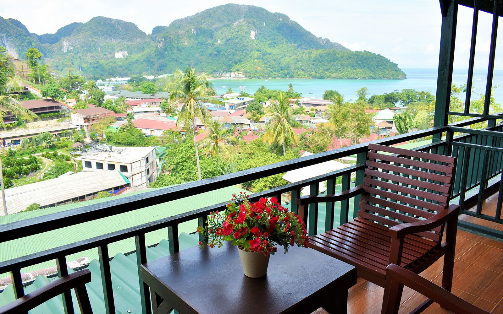A balcony with a wooden chair and a table, a potted plant, and a view of a coastal town with mountains in the background.