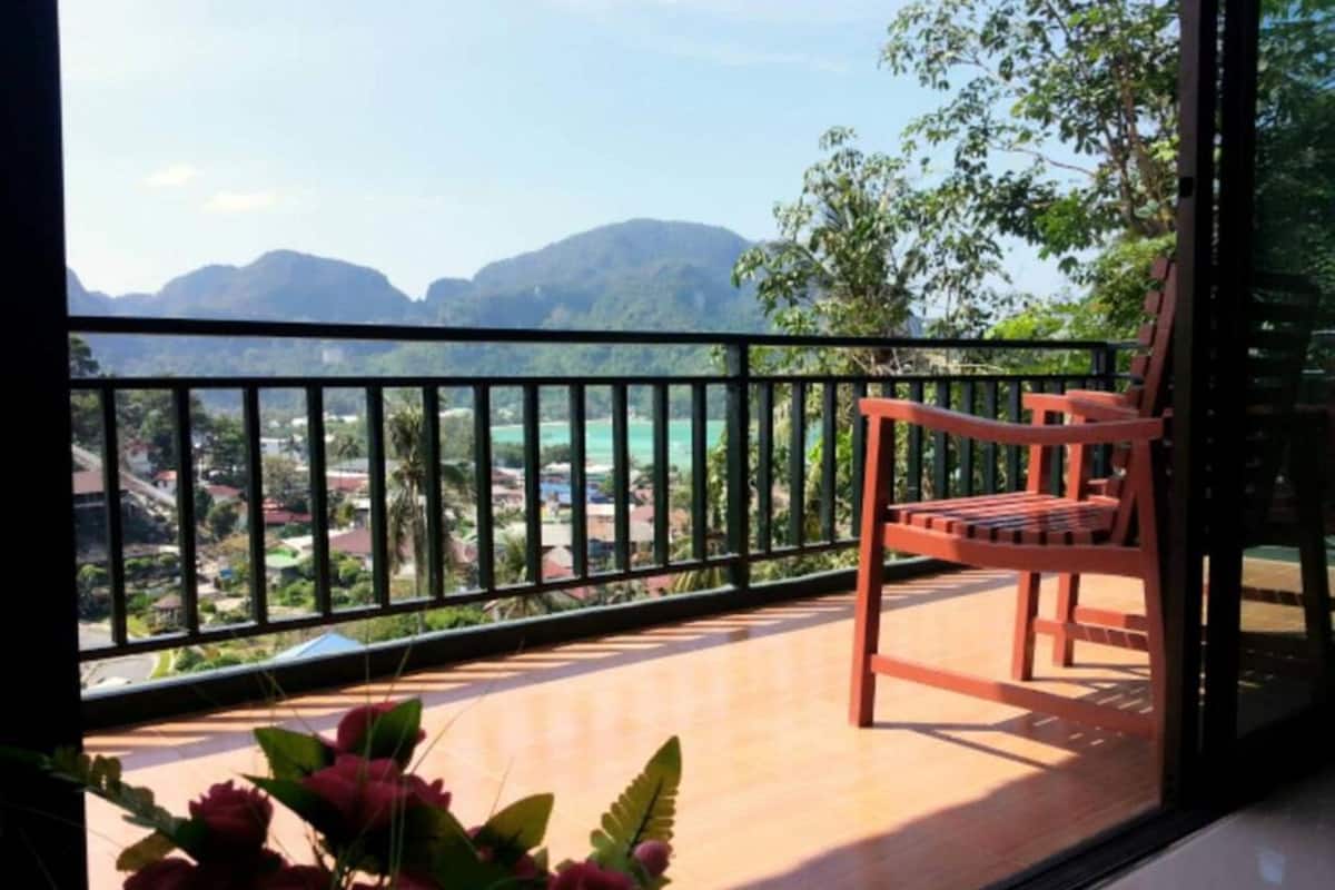 Un balcón con una silla de madera, vista a un paisaje montañoso y una barandilla.