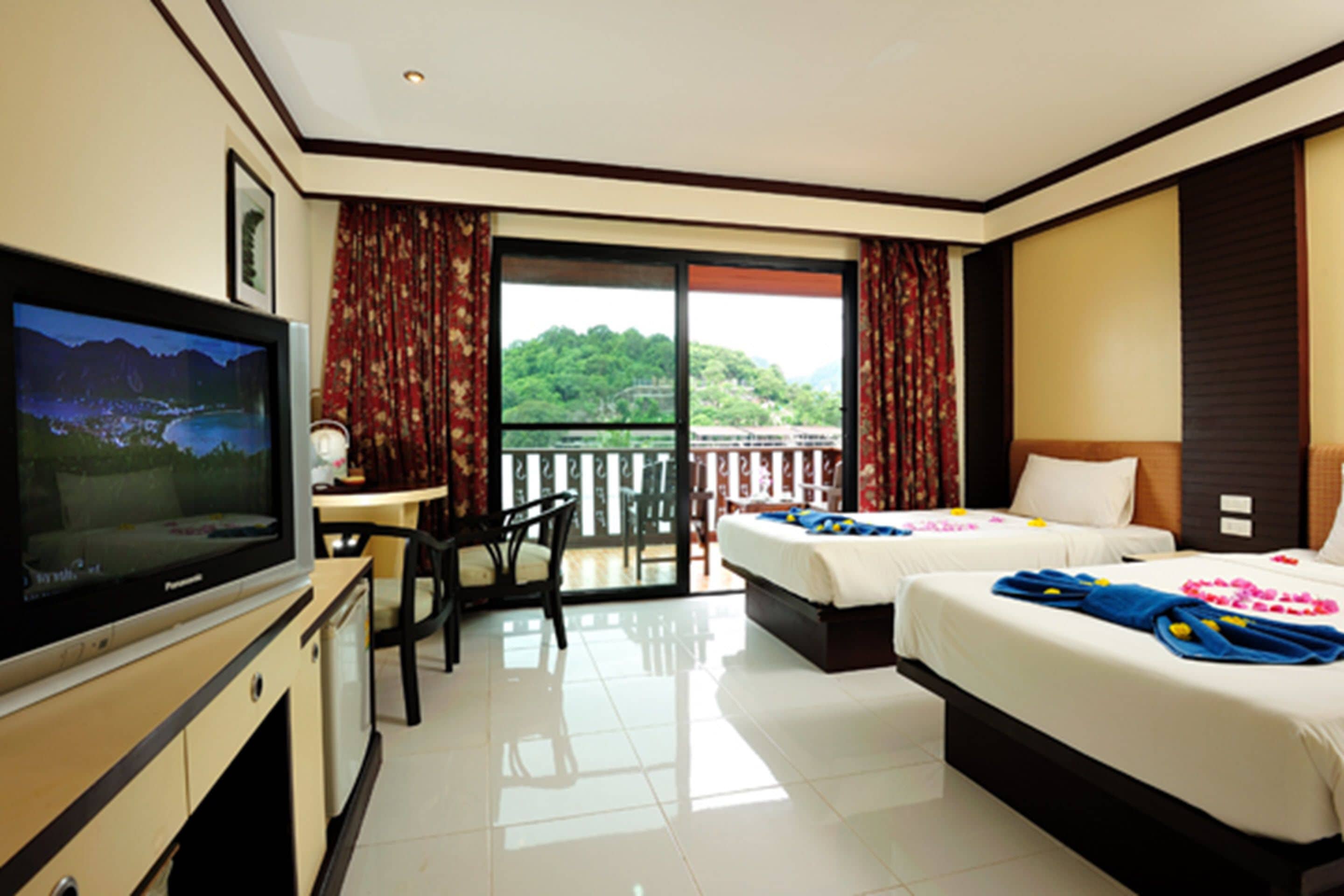 A hotel room with a TV, two beds, a desk, and a balcony with a scenic view.
