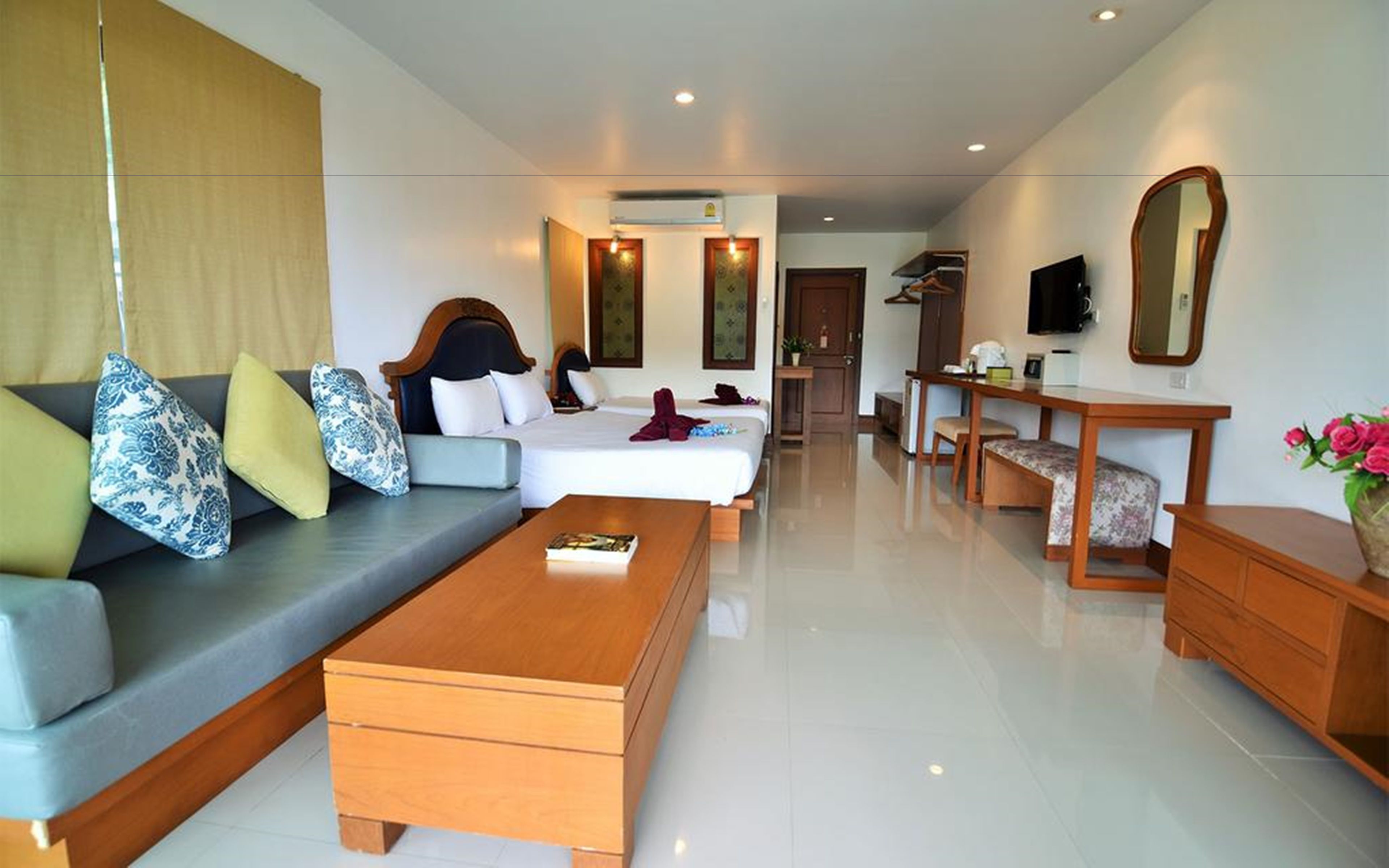 A modern hotel room with a sofa, two beds, a wooden coffee table, a desk with a chair, a TV, and a mirror.