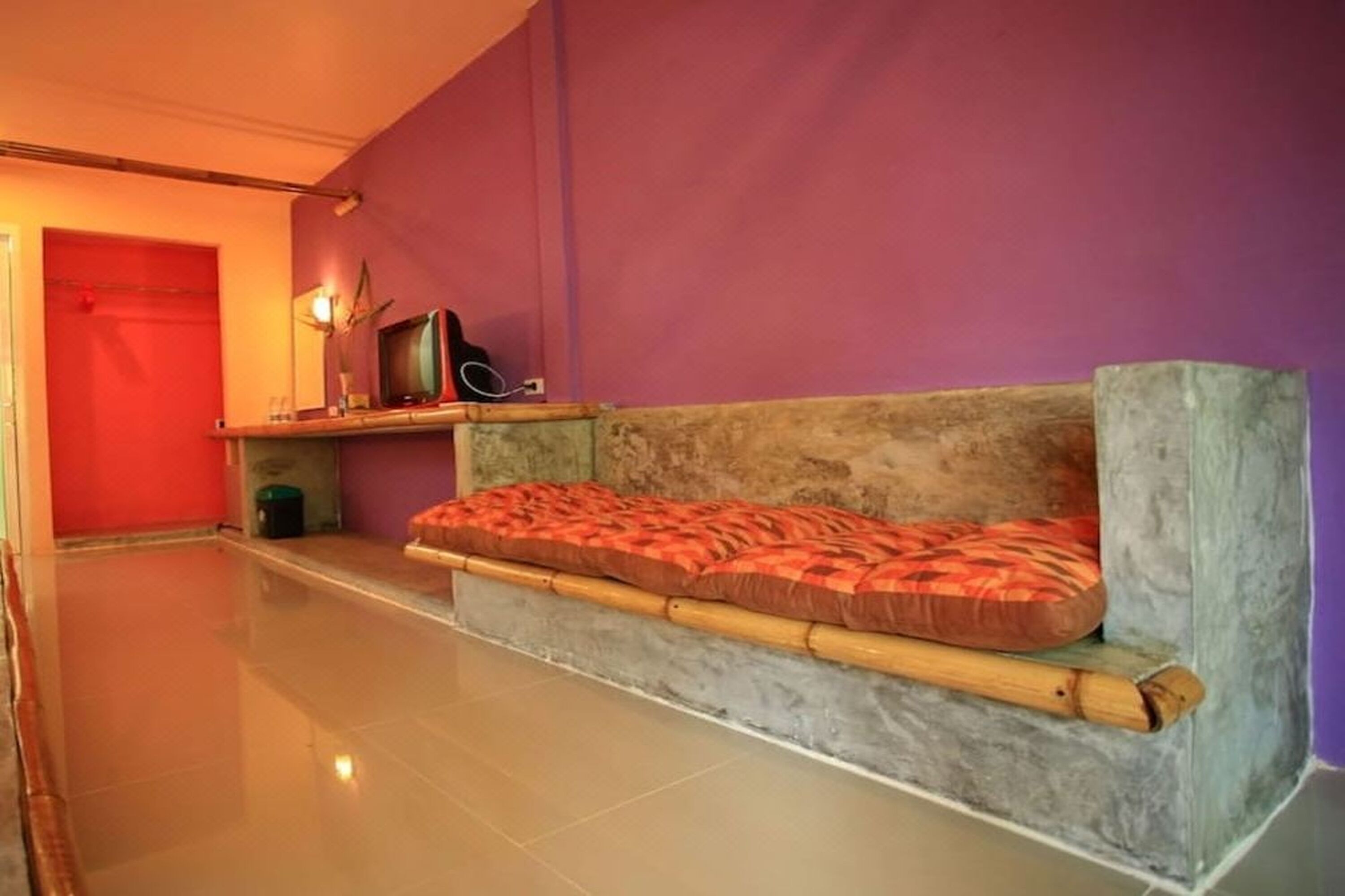 A room with a low, built-in bench with a red patterned cushion, a television on a shelf, and a red door in the background.