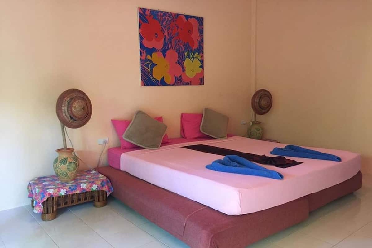 A bedroom with a bed, pillows, a small table with a vase, and a wall painting.