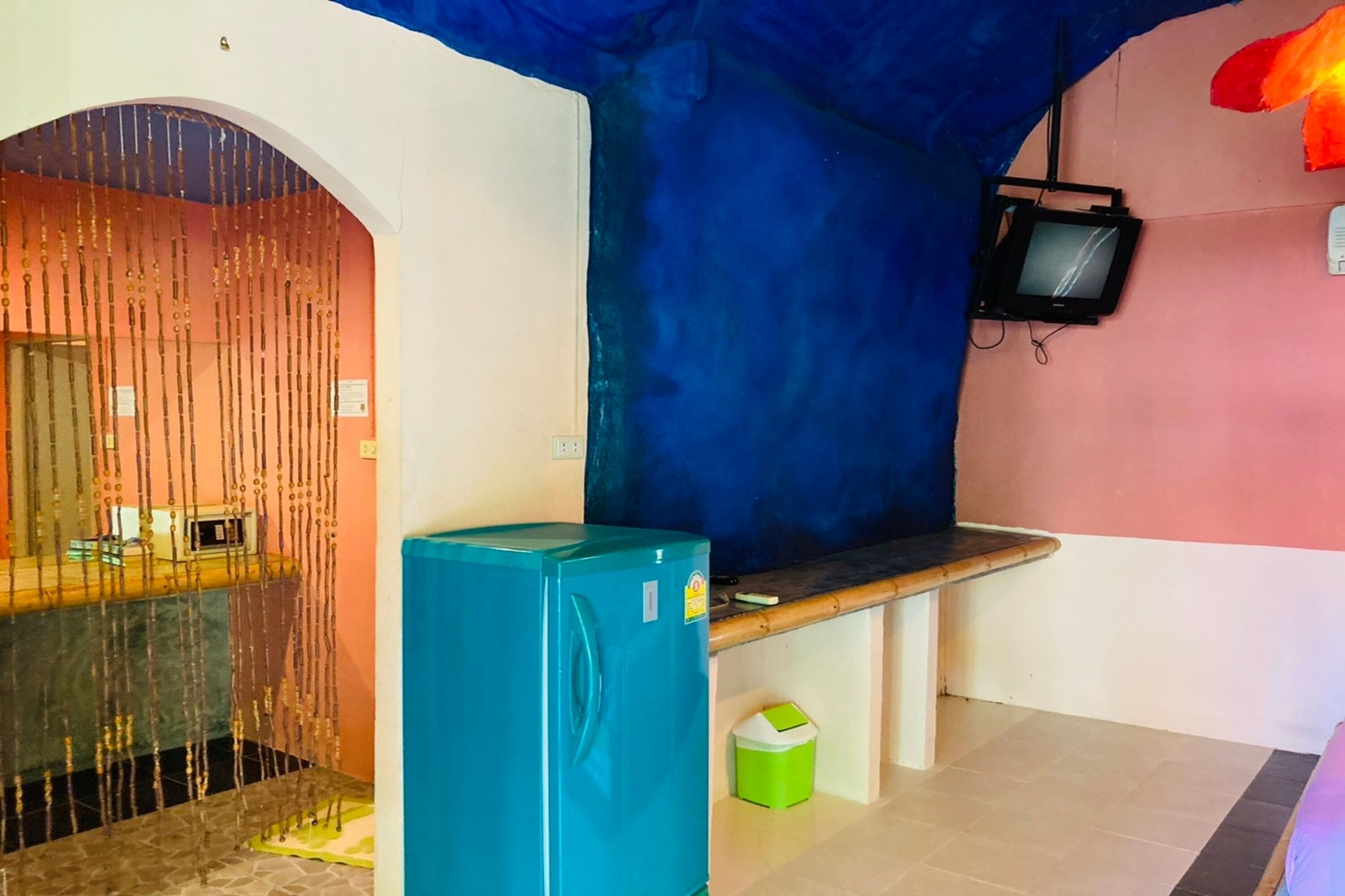A room with a blue refrigerator, a bench, a television mounted on the wall, and a door with a decorative curtain.