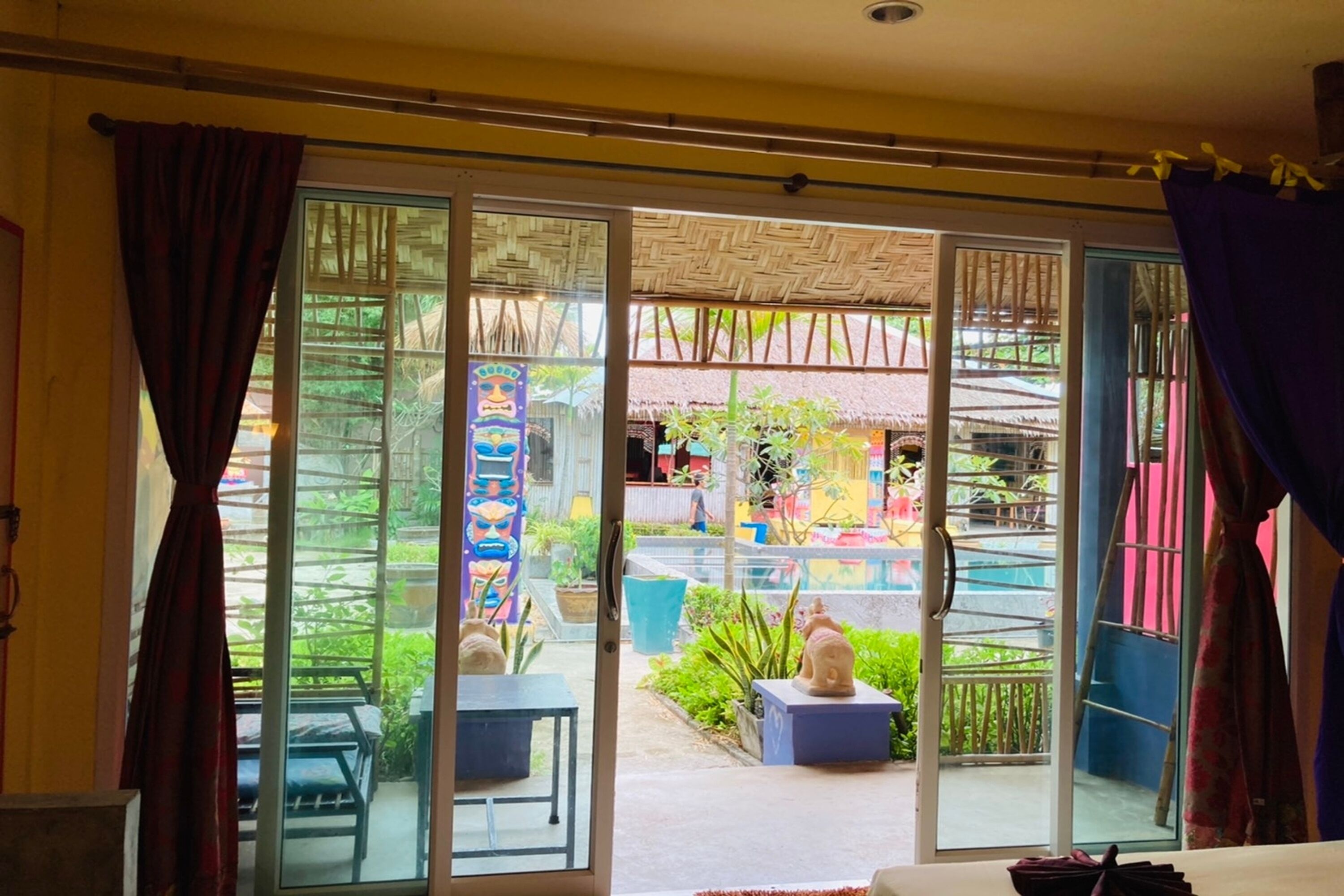 A room with a sliding glass door leading to an outdoor area with a thatched roof, potted plants, and a statue.