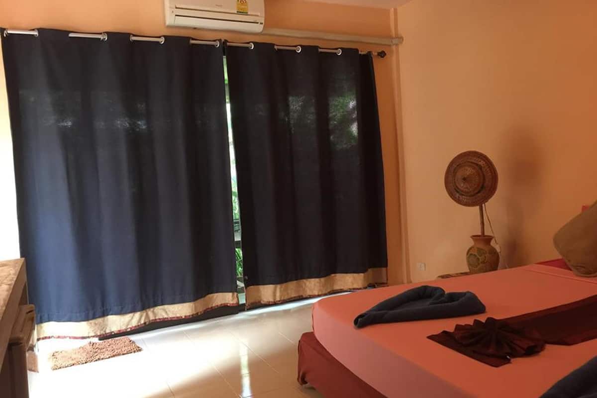 A bedroom with a bed, an air conditioner, a window with curtains, and a small table with a vase.