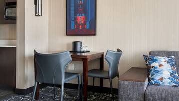 A hotel room with a dining area featuring a wooden table, two chairs, a small coffee table, and a couch with a patterned cushion. A framed artwork of a building is on the wall.