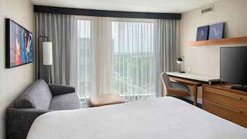 Ein Hotelzimmer mit einem großen Bett, einem Schreibtisch, einem Sessel und einem Fernseher. Es gibt ein Fenster mit Vorhängen und einen Blick auf Grünfläche außerhalb.