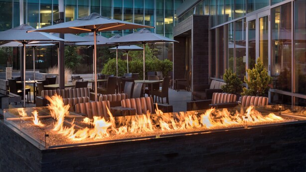 An outdoor seating area with a fire feature, tables, chairs, and umbrellas. - Courtyard Philadelphia South at The Navy Yard (Philadelphia)