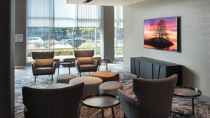 A modern lounge area with comfortable seating, a large window with a view of parked cars, and a wall-mounted TV. - Courtyard Philadelphia South at The Navy Yard (Philadelphia)