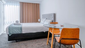 A hotel room with a bed, a round glass table, two orange chairs, a grey headboard, and a window with curtains. - Merivale Court Motel (Christchurch)