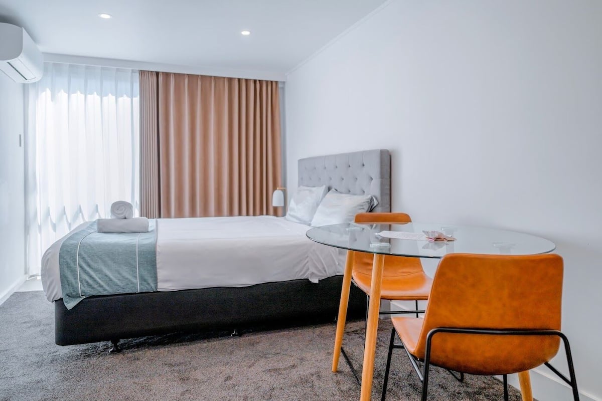 A hotel room with a bed, a round glass table, two orange chairs, a grey headboard, and a window with curtains.