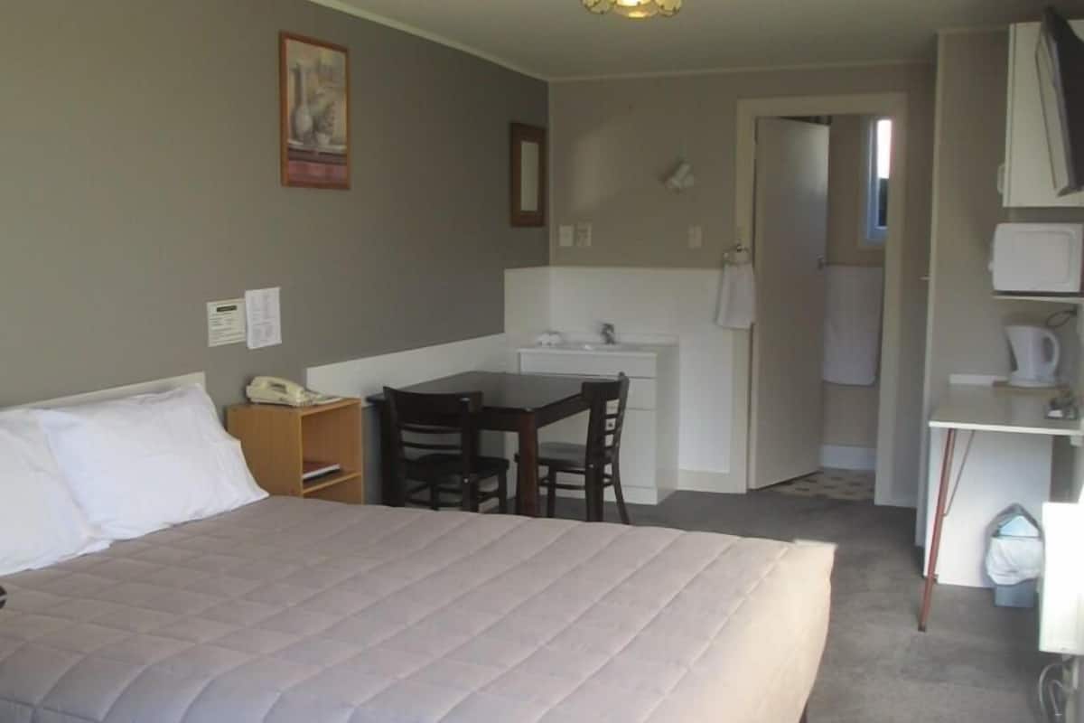A hotel room with a large bed, a desk, a chair, a small kitchenette, and a bathroom visible through an open door.