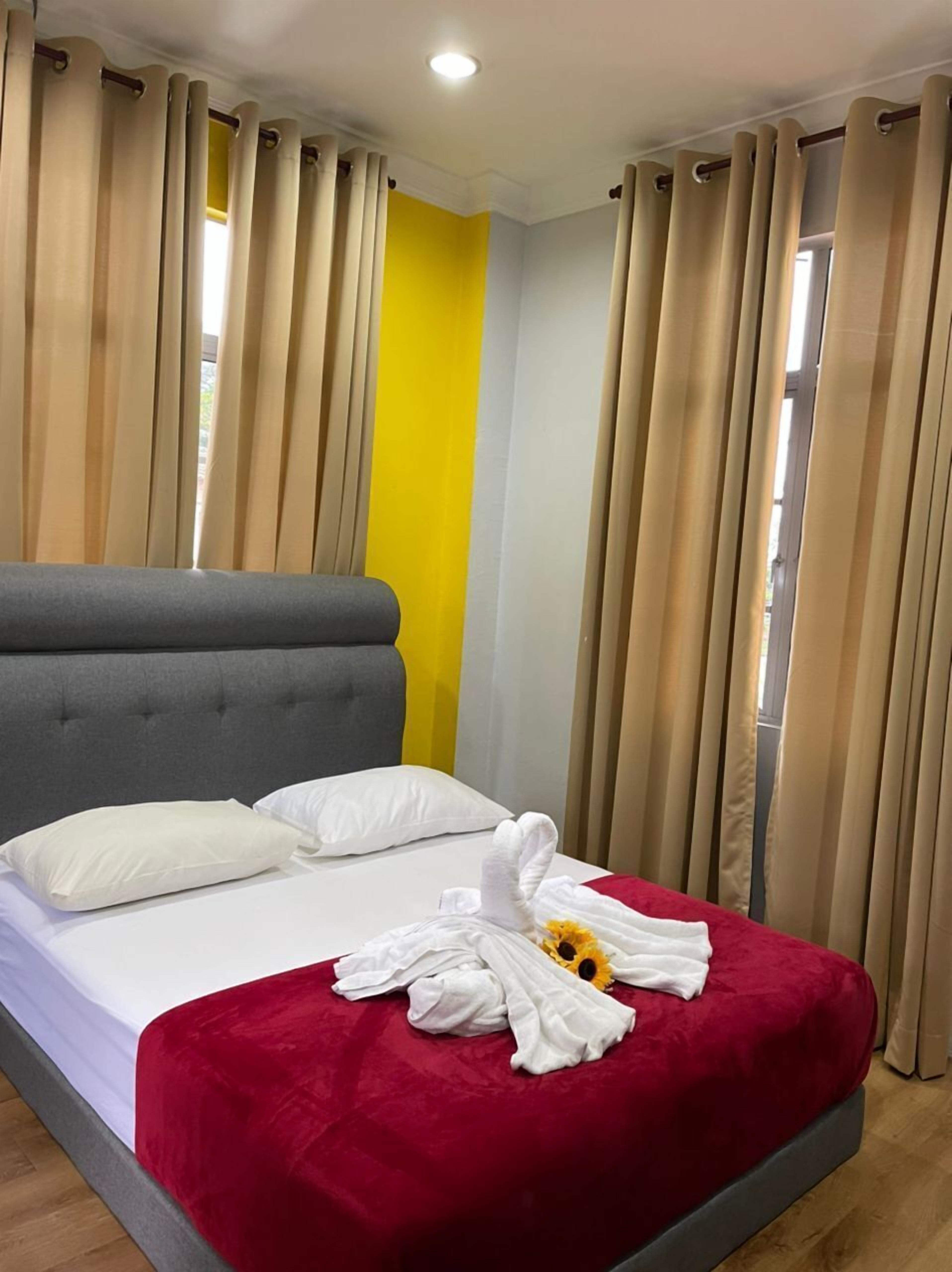 Ein Hotelzimmer mit Bett, beigen Vorhängen, einer roten Bettdecke und einem weißen Handtuch mit einem Sonnenblumenmuster.