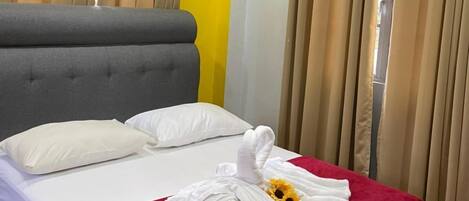 Habitación de hotel con cama, cortinas color beige, colcha roja y toalla blanca con un girasol.