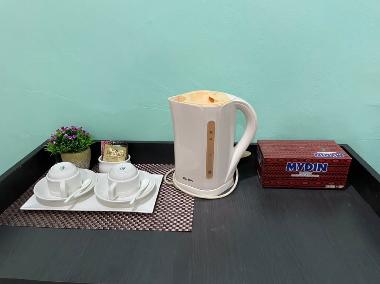 Un plan de travail de cuisine comprenant une bouilloire électrique blanche, deux tasses à thé blanches avec leurs soucoupes, une petite plante en pot, un paquet de thé et une boîte de thé MyDIN.