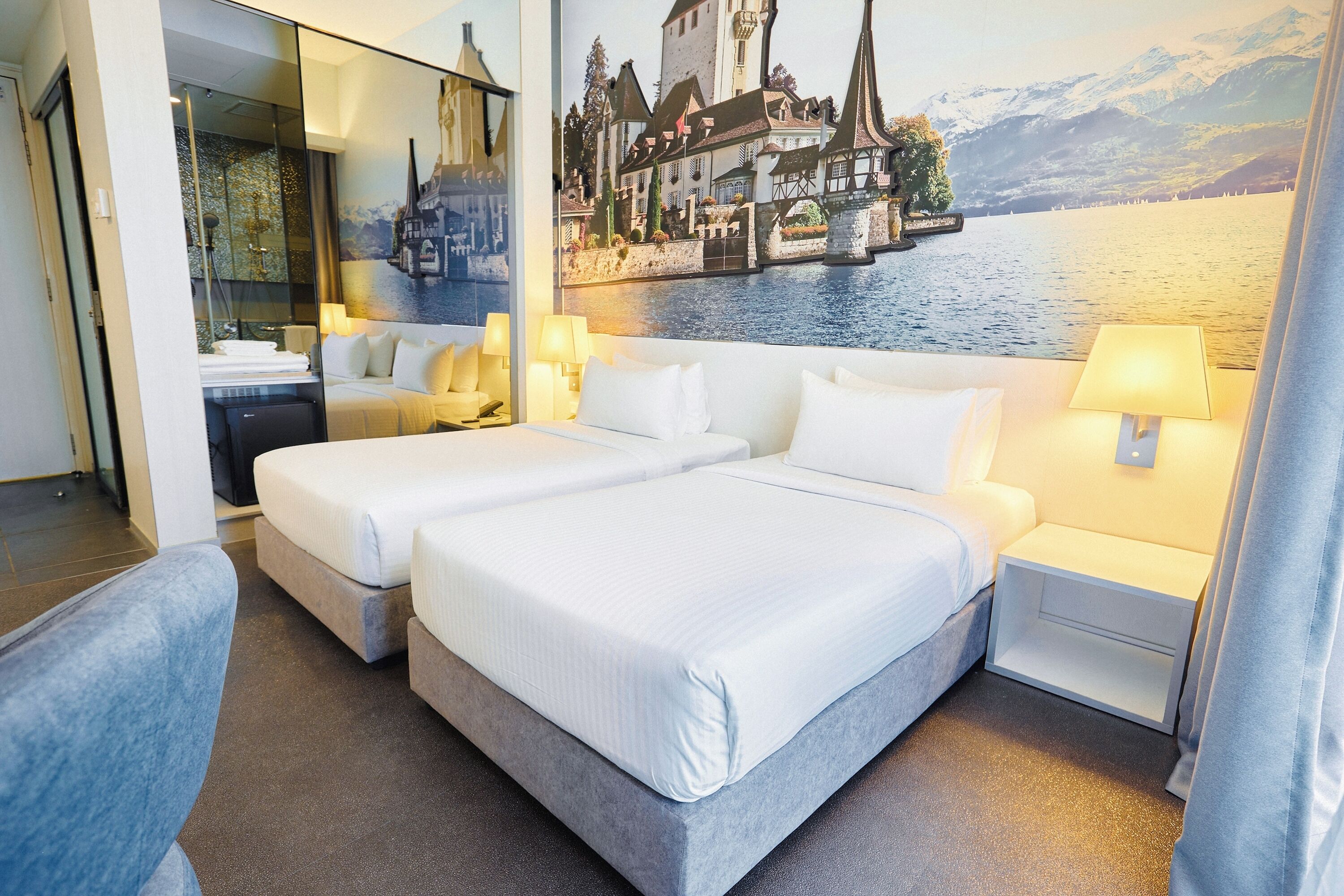 A hotel room with two beds, a large mirror, and a scenic mural of a lakeside town.