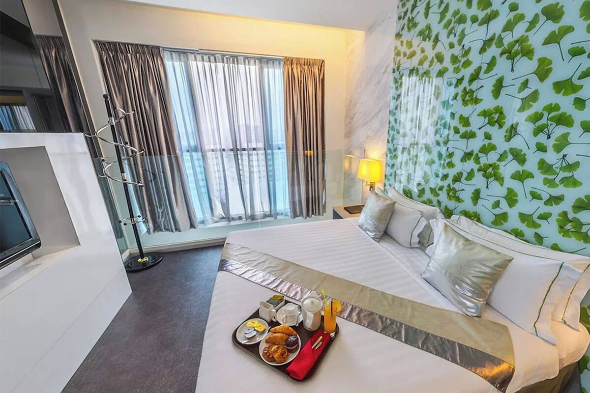 Ein Hotelzimmer mit Bett, Frühstückswagen, Fernseher, Fenster mit Vorhängen und einer Wand mit Blättermotiv.
