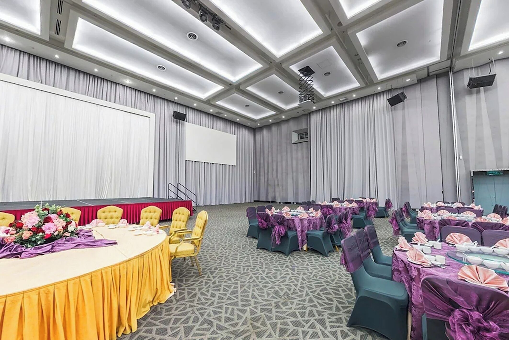 Une salle de conférence équipée d’une scène, de rangées de tables avec des chaises et d’un grand écran.