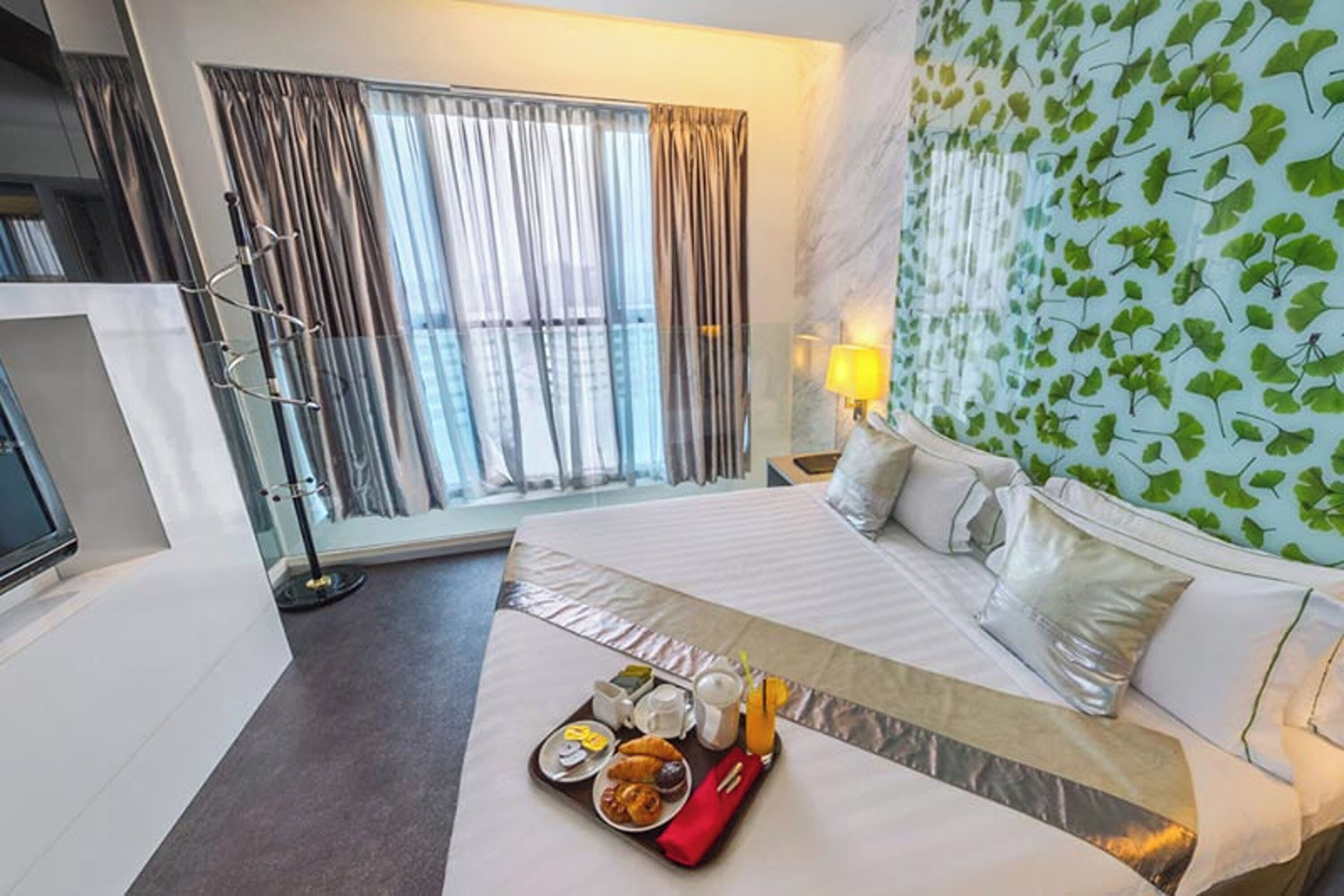 Une chambre d’hôtel avec un lit, une fenêtre avec des rideaux, une table de chevet avec une lampe et un plateau contenant des articles pour le petit-déjeuner.