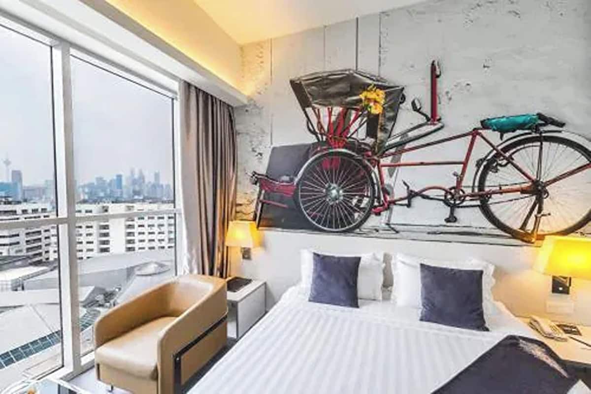 Ein Hotelzimmer mit großem Fenster, einem Bett mit weißer Bettwäsche, zwei Kissen, einem Sessel und einem an der Wand befestigten Fahrrad.