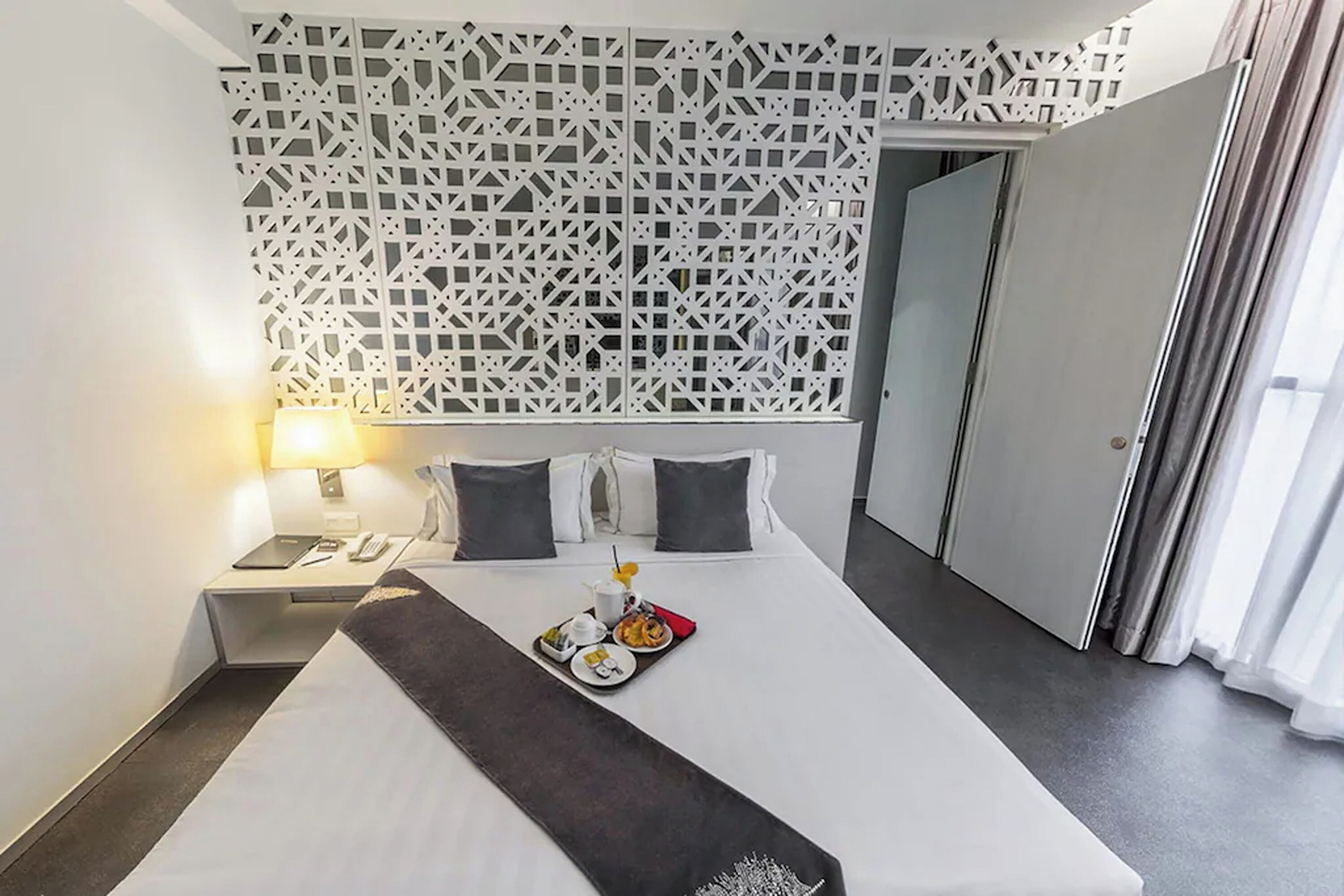 Habitación de hotel con una cama bien hecha, una bandeja con alimentos para el desayuno, una lámpara de noche y un panel decorativo en la pared.