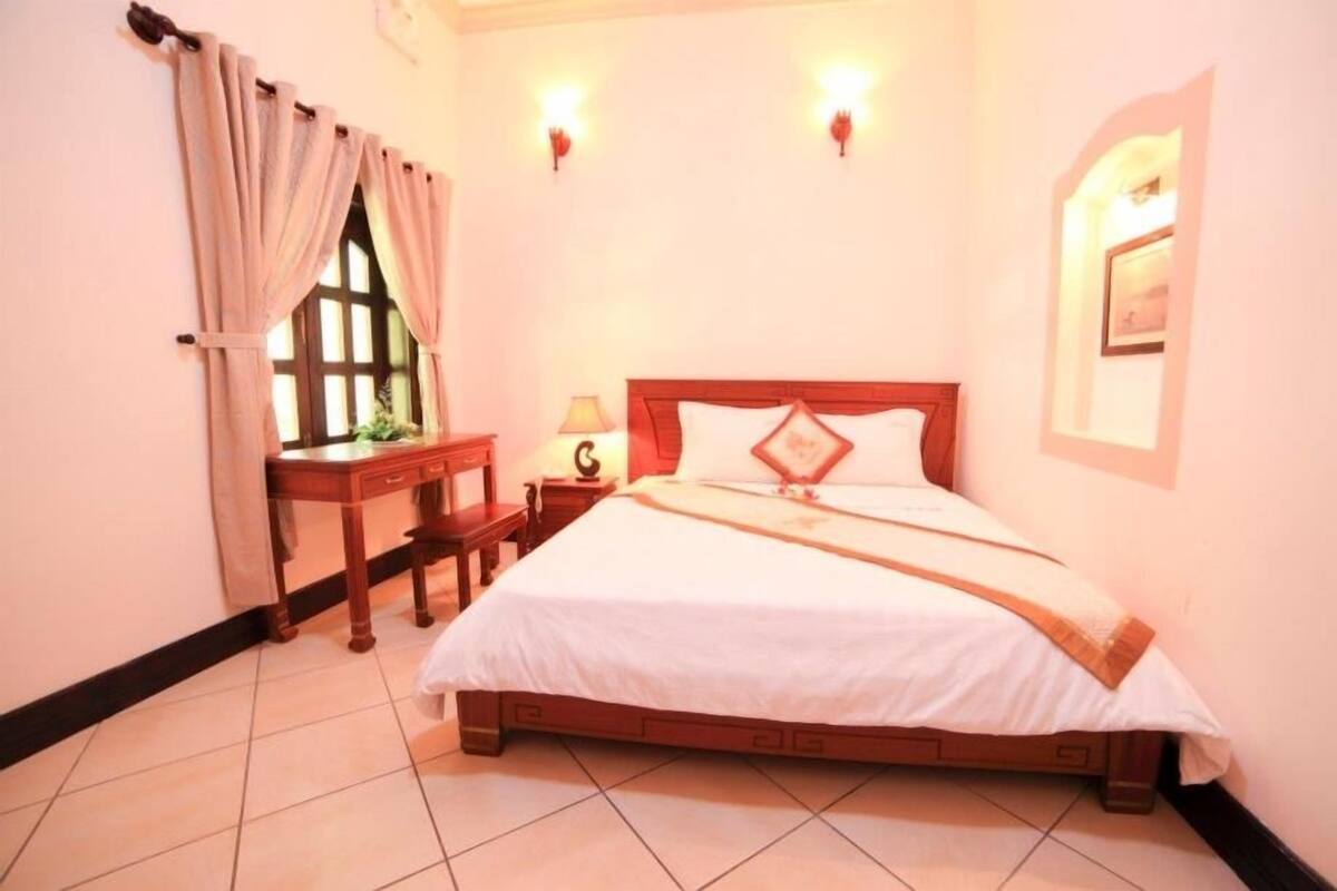 A bedroom with a wooden bed, a nightstand with a lamp, a window with curtains, and wall-mounted lights.