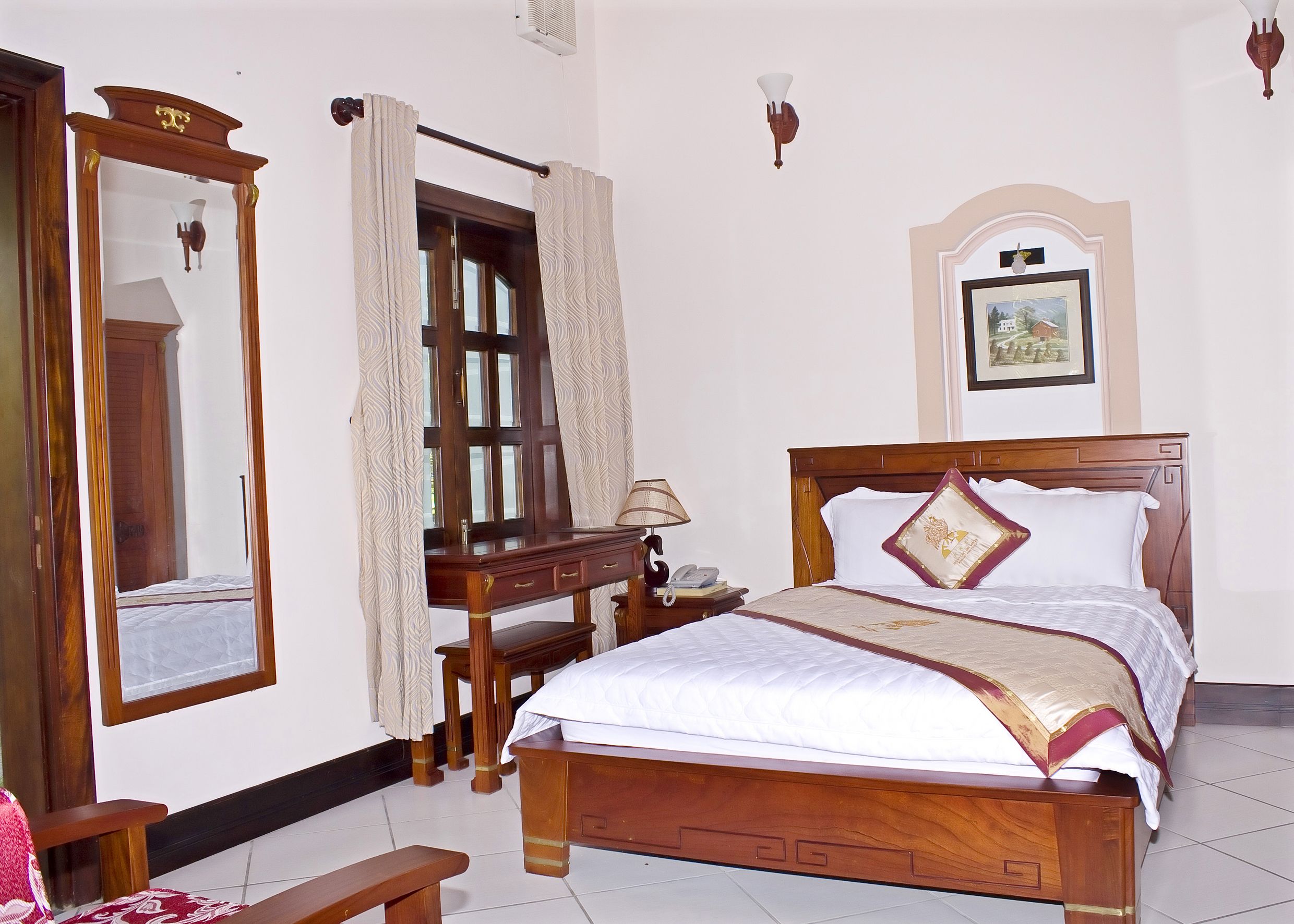 A neatly arranged bedroom with a wooden bed, a bedside table with a lamp, a mirror, and a framed picture on the wall.
