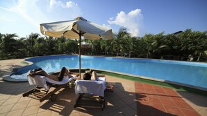 Zona junto a la piscina con sillones de descanso y sombrillas, rodeada de palmeras y un cielo despejado.