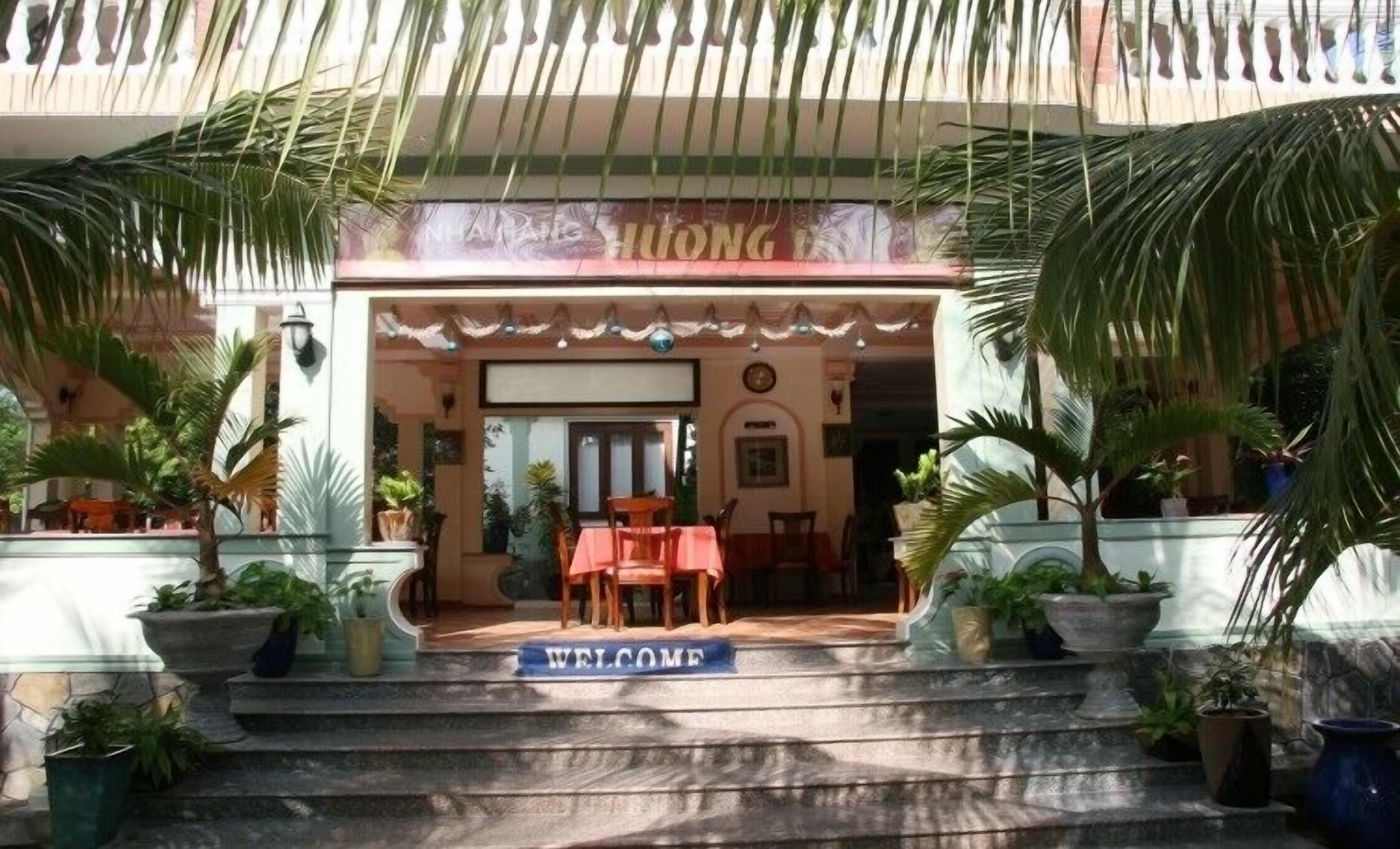 Entrada de hotel con un letrero que dice 'WELCOME', palmeras y una zona de estar cubierta.