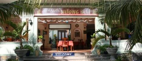 Entrada de hotel con un letrero que dice 'WELCOME', palmeras y una zona de estar cubierta.
