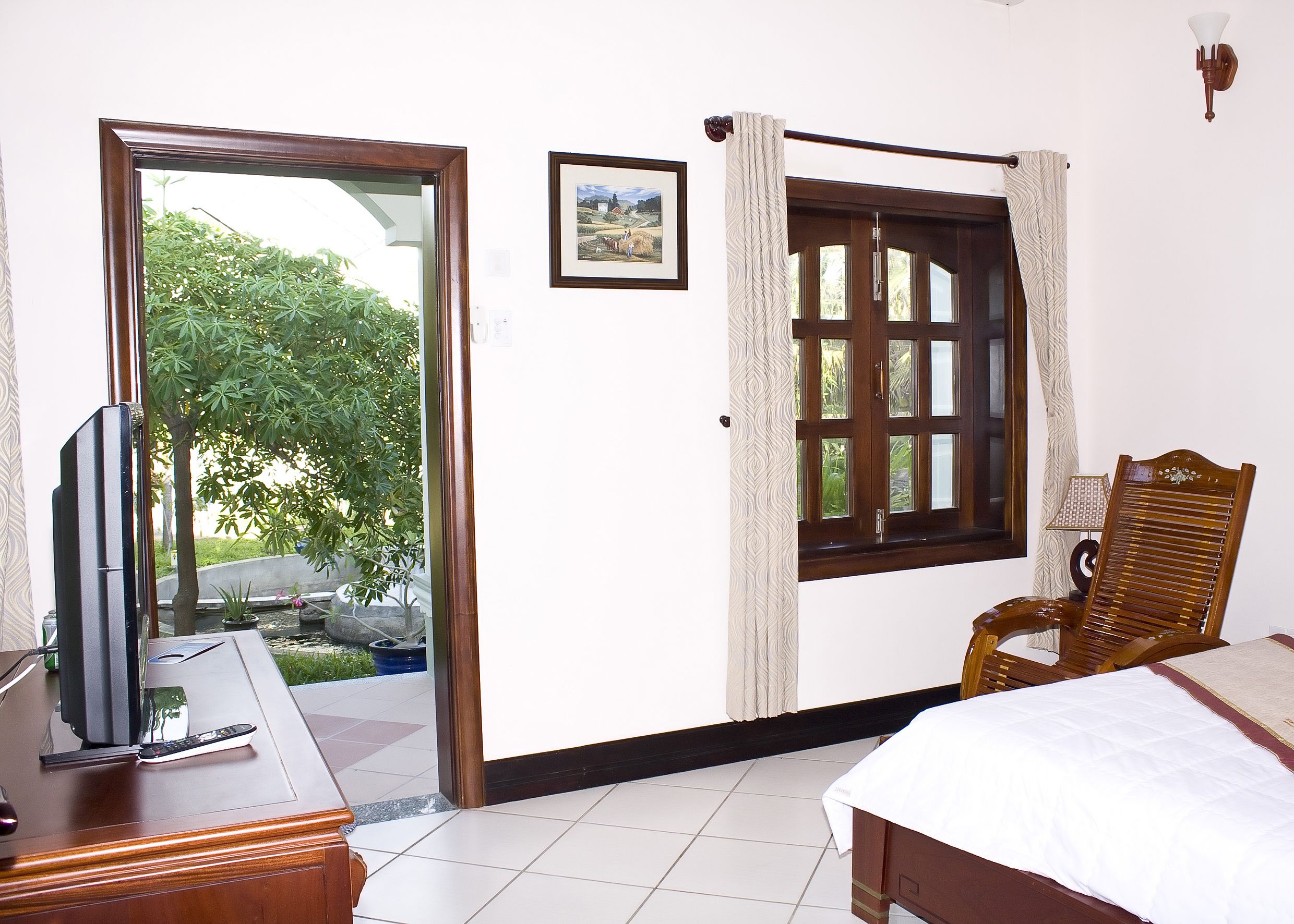 Habitación de hotel con cama, una silla de madera, escritorio con televisor y control remoto, ventana con cortinas y un cuadro en la pared.