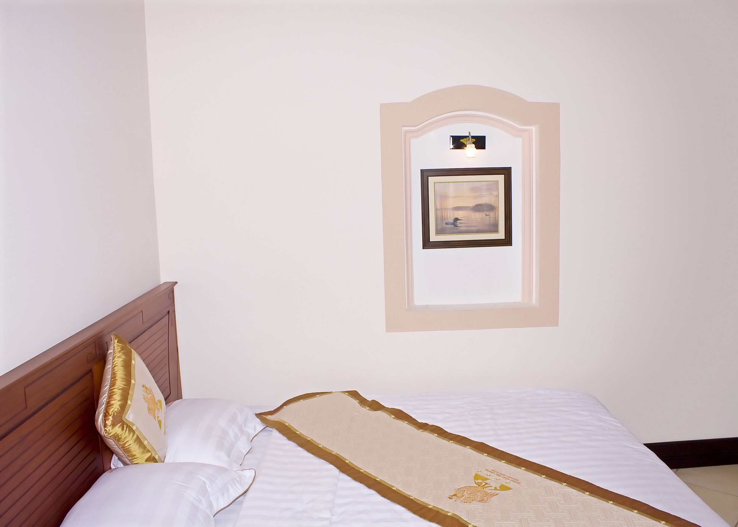 A neatly made bed with a white and gold bedspread, a wooden headboard, and a framed picture on the wall.