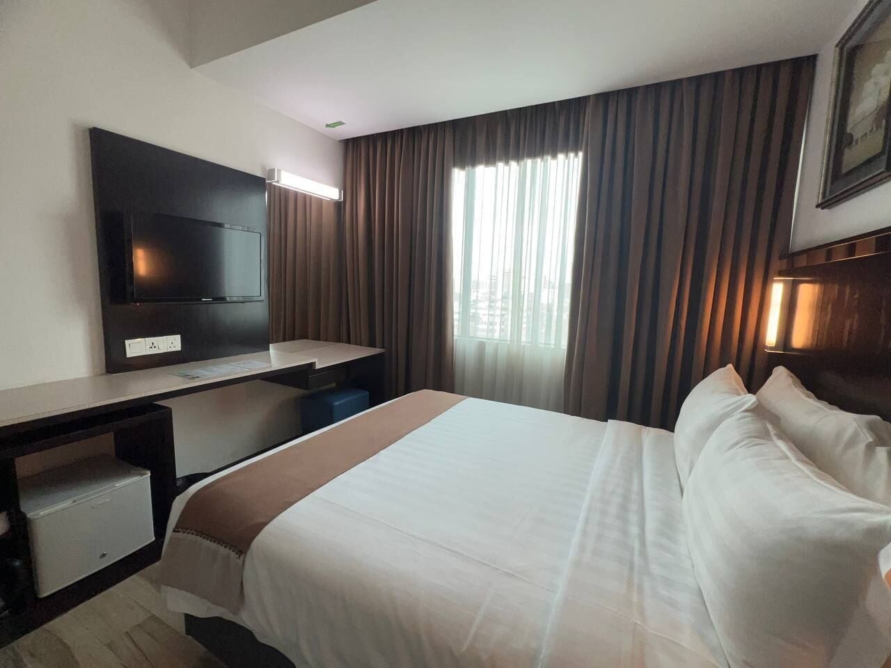Una habitación de hotel con cama, escritorio, televisor y ventana con cortinas.
