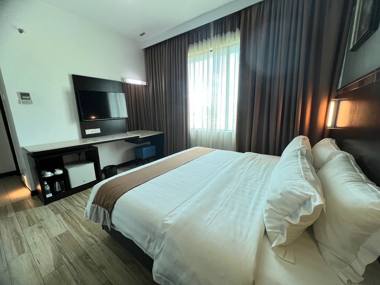 A hotel room with a large bed, a TV mounted on the wall, a desk with a computer, and a window with curtains.