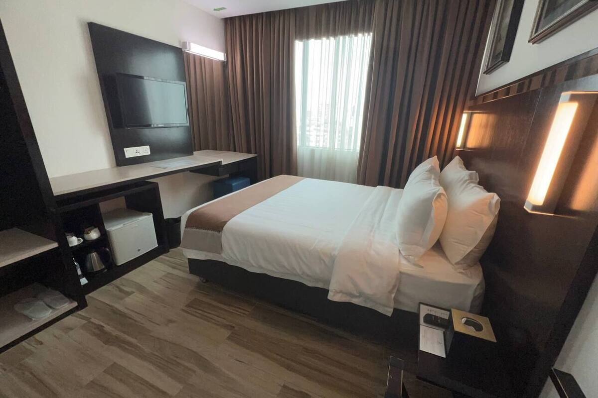 A hotel room with a bed, a TV mounted on the wall, a desk with a chair, and a window with curtains.