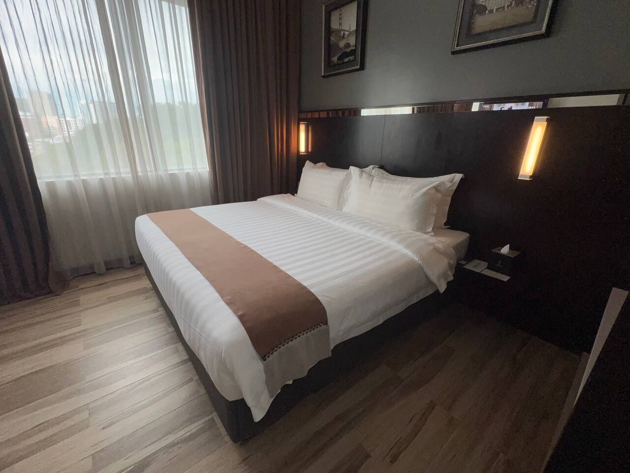 A hotel room with a large bed, wooden flooring, a headboard with integrated lighting, and a window with sheer curtains.
