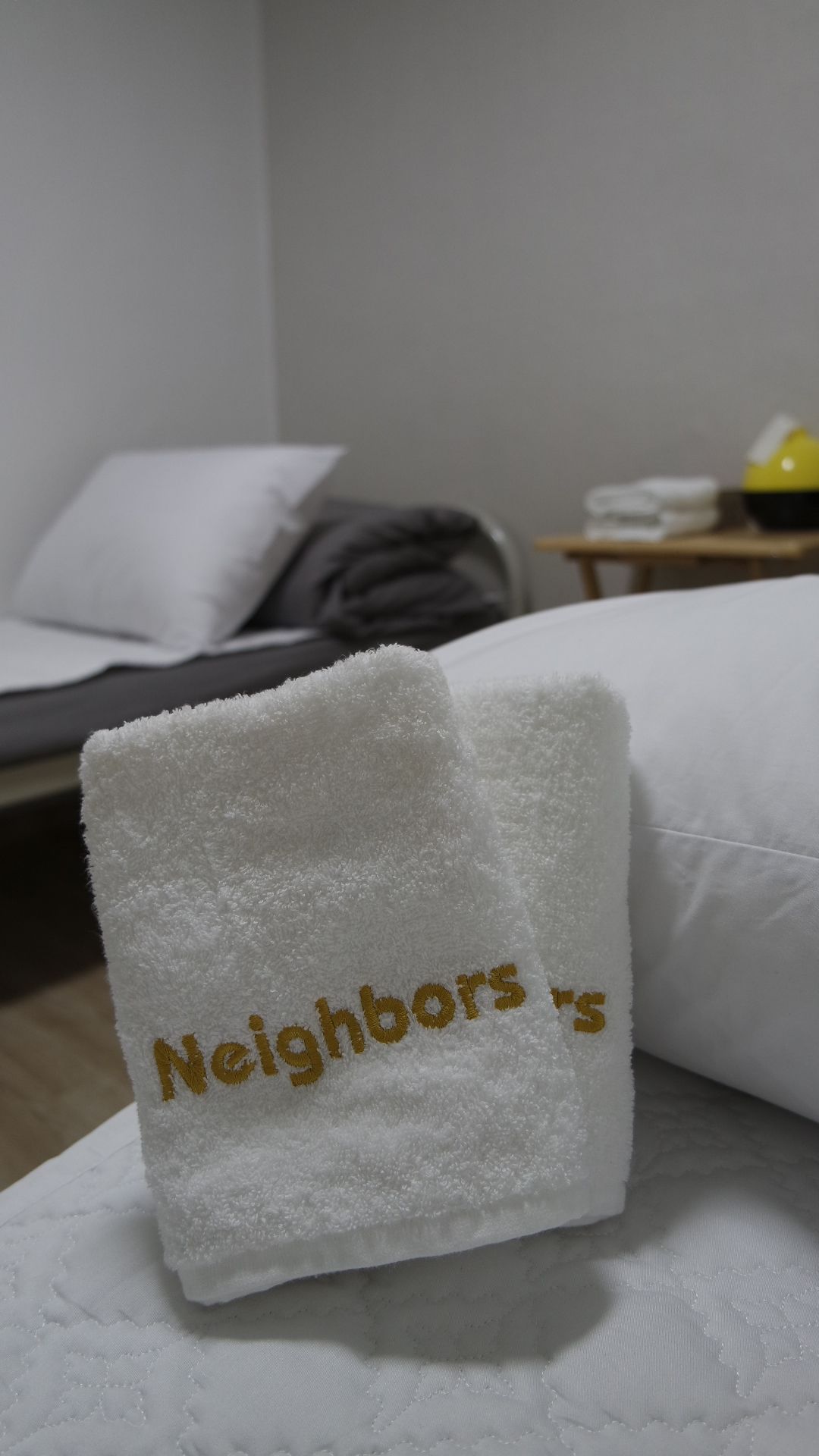 A neatly made bed with a white pillow and a folded white towel with "Neighbors" embroidered on it.
