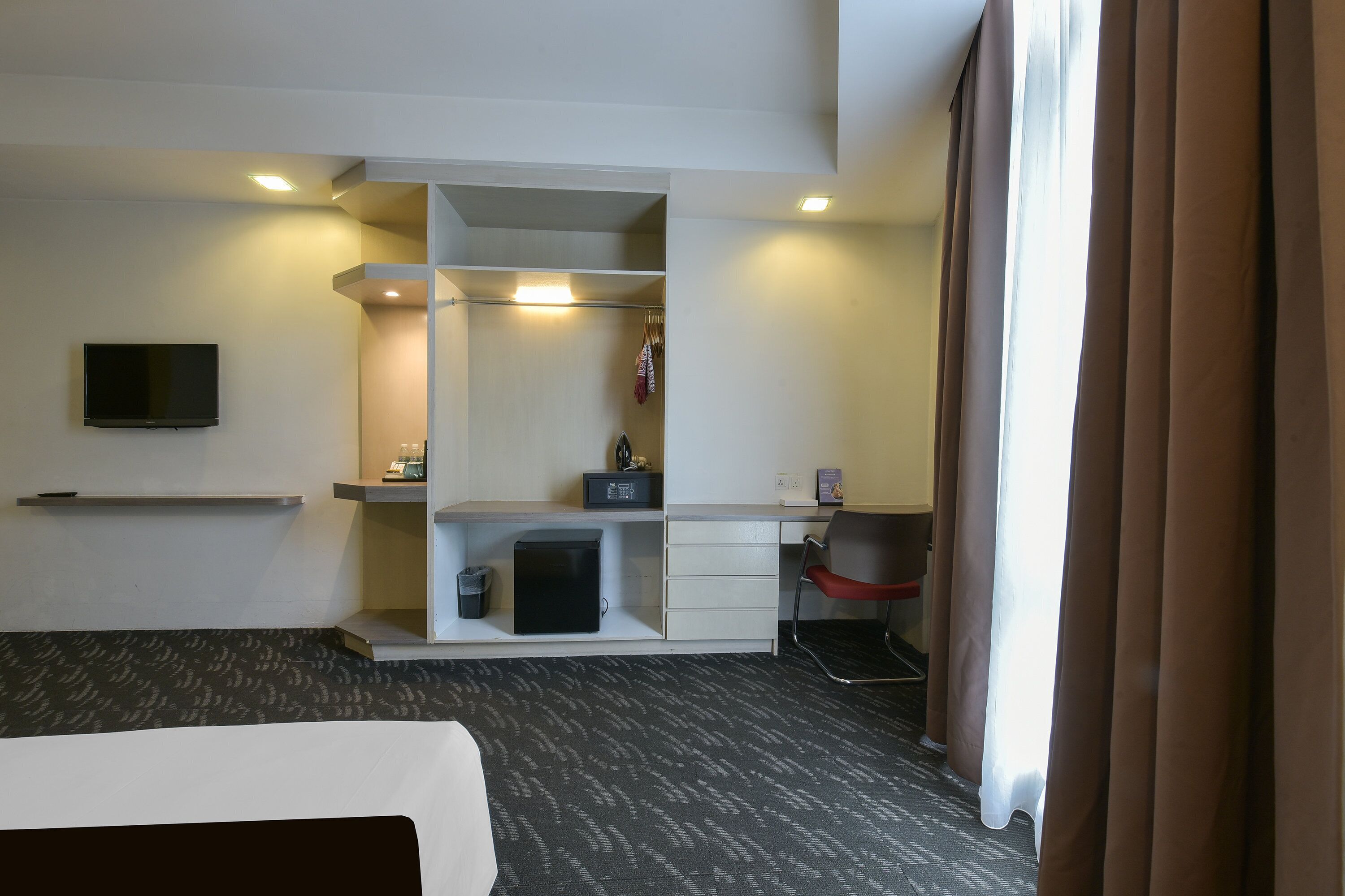Habitación de hotel con cama, escritorio, silla, televisor montado en la pared y armario.