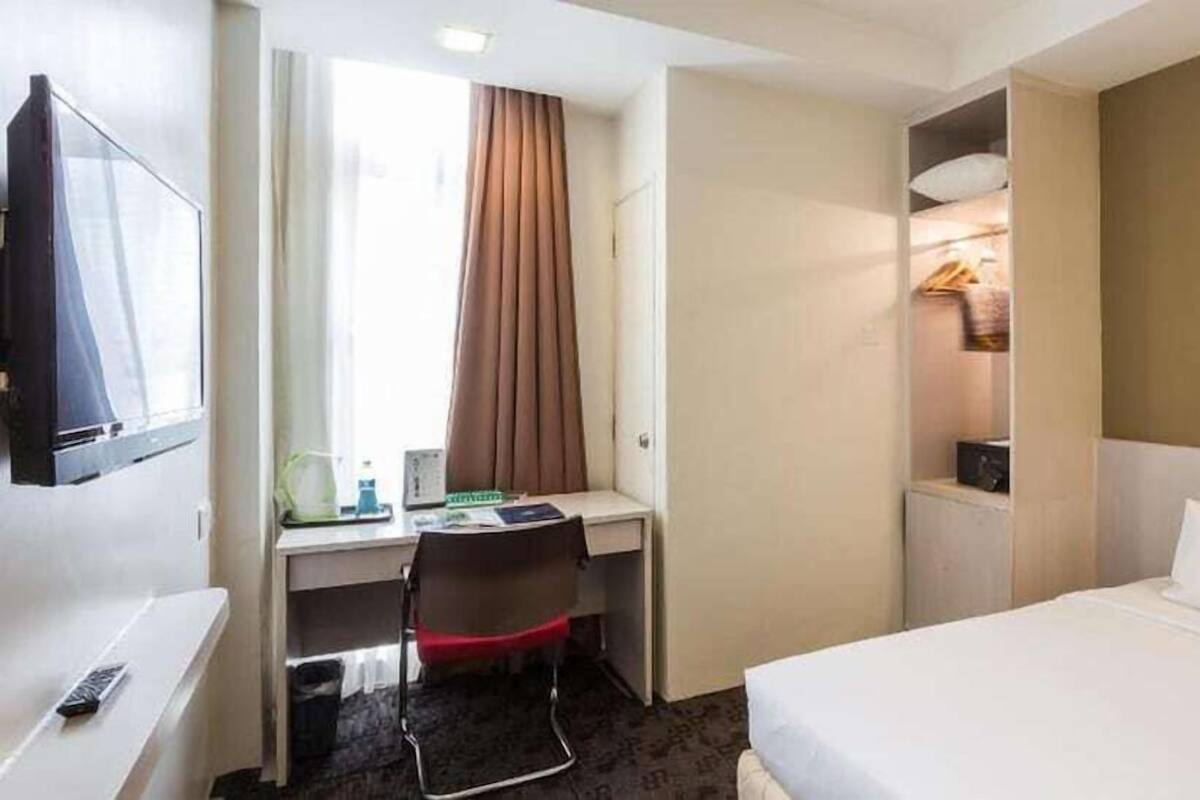 A hotel room with a bed, a desk with a chair, a television, a window with curtains, and a shelf with items.