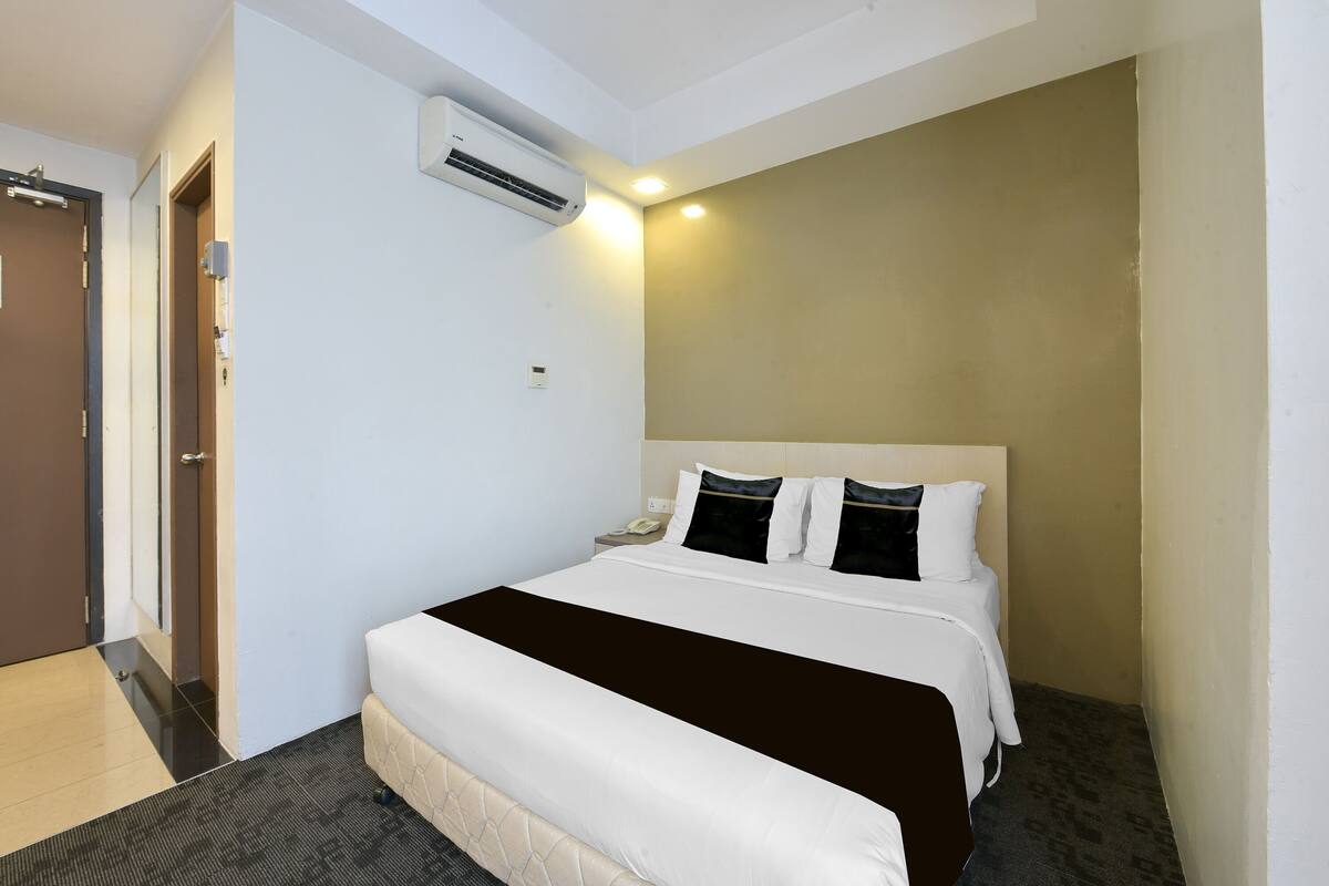 A hotel room with a bed, a bedside table, an air conditioning unit, and a door.