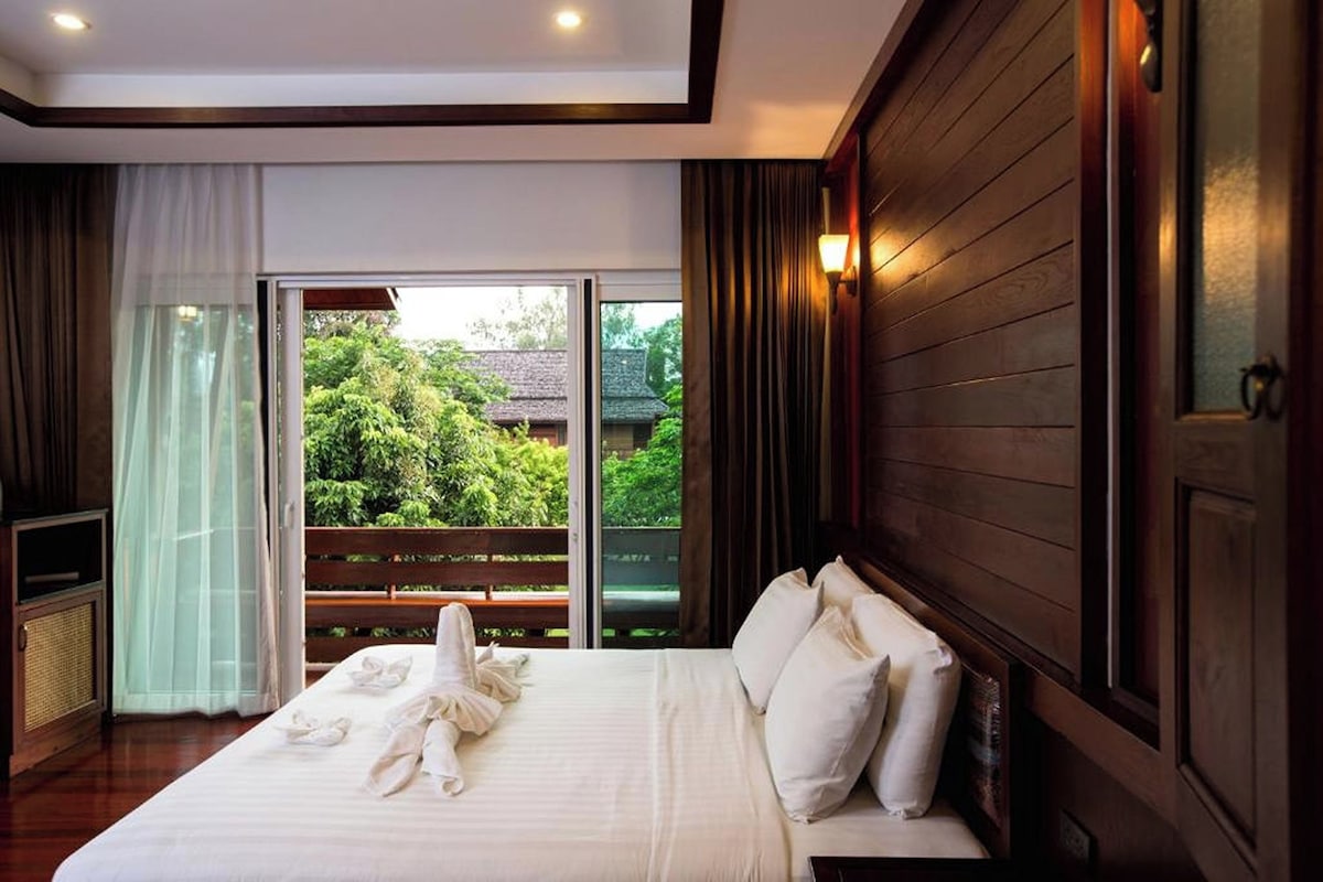 A hotel room with a bed, a window with curtains, and a view of greenery outside.