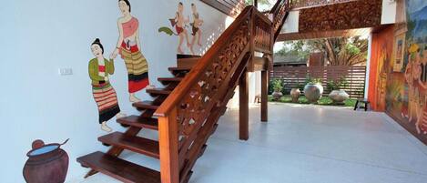 A staircase with intricate wooden railings leading to a balcony, a mural depicting traditional figures on the wall, and a tiled floor.