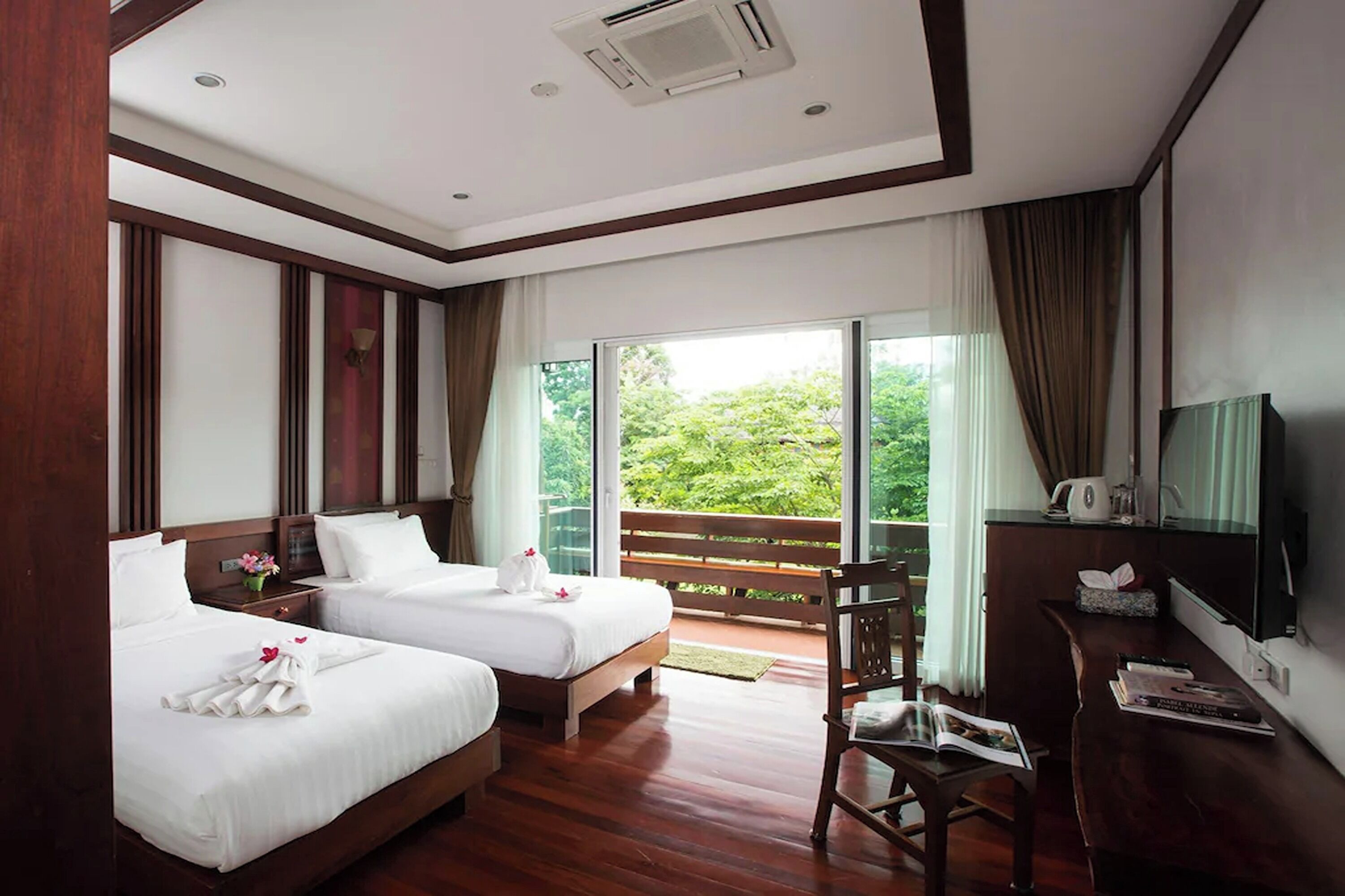 A hotel room with two beds, a desk, a chair, a TV, and a balcony with a view of greenery.