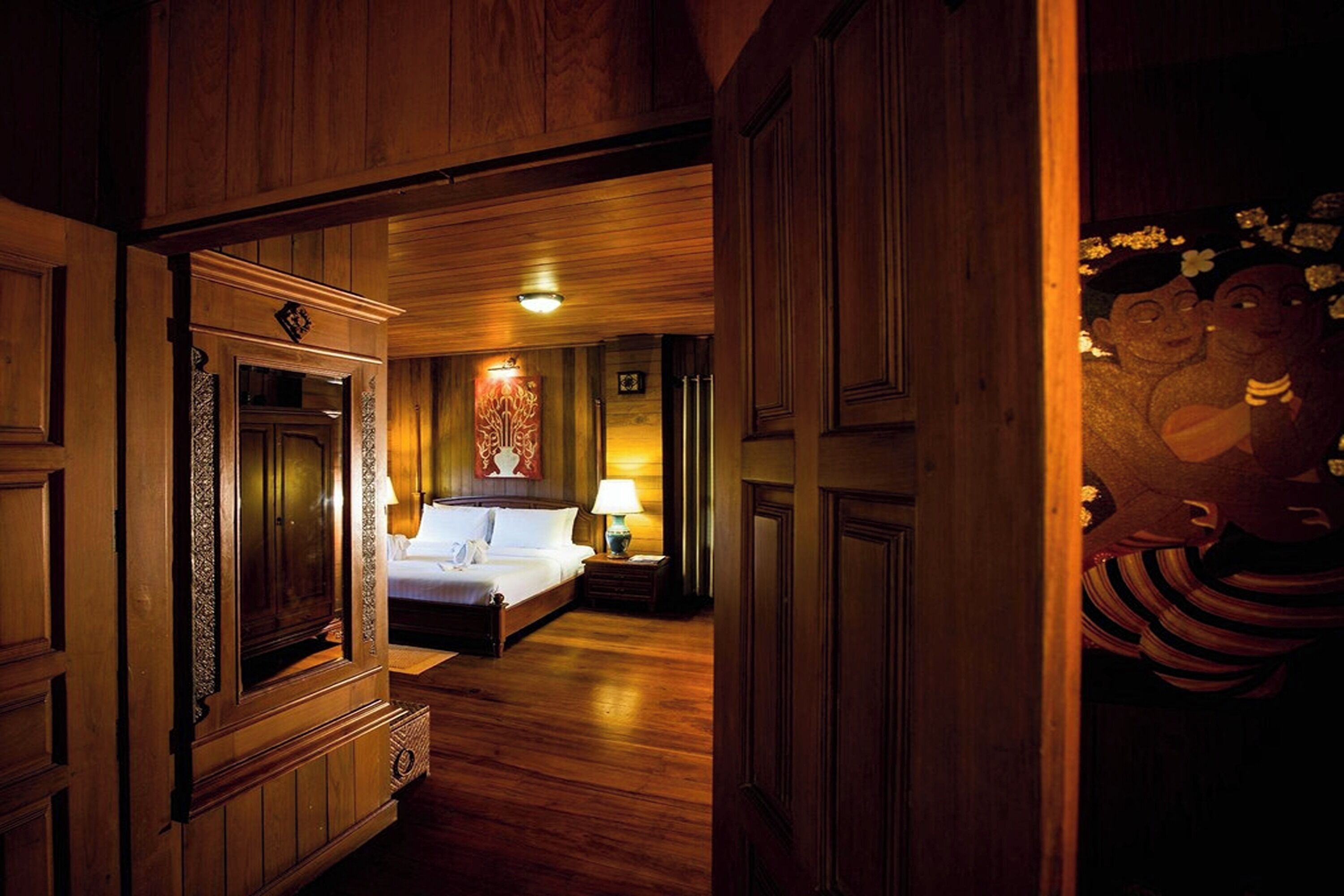 A wooden-floored hotel room with a bed, a nightstand, and a wall-mounted lamp.