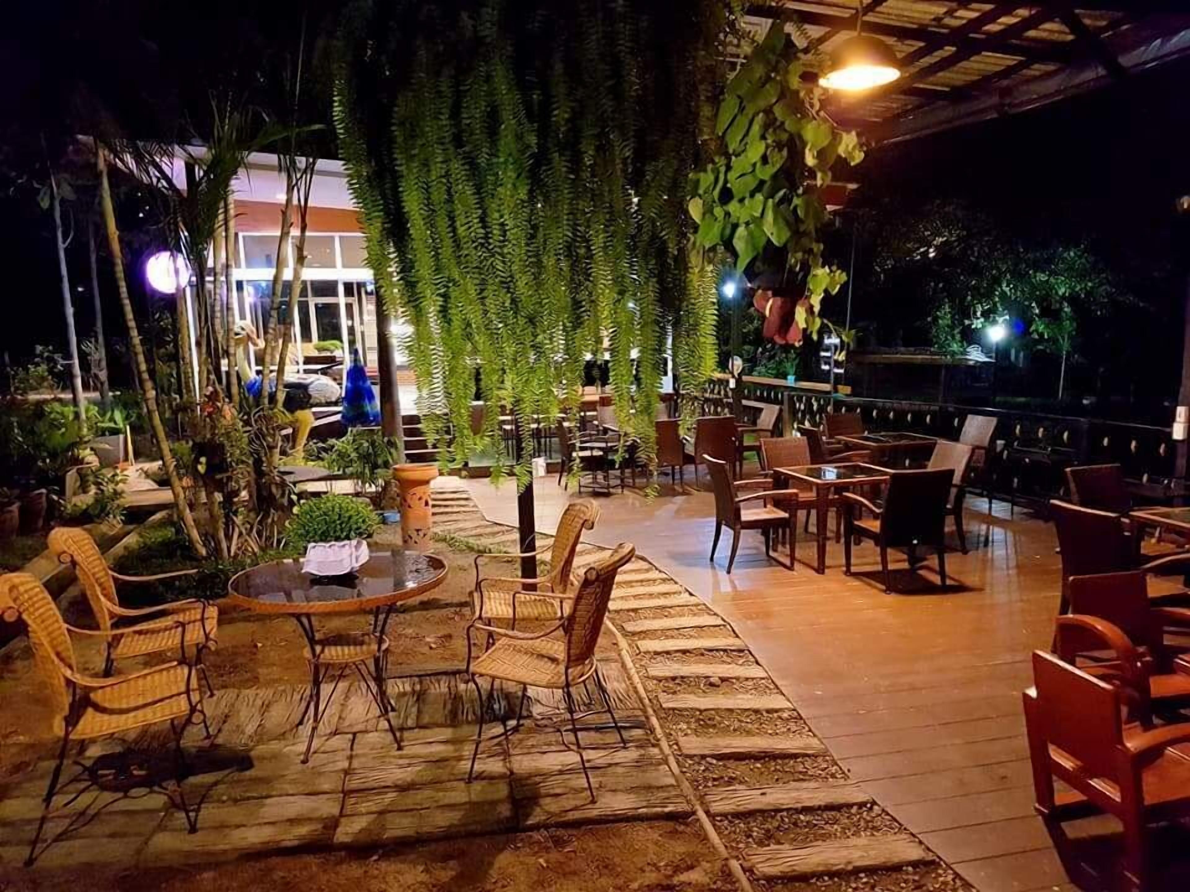 An outdoor patio area with wooden tables and chairs, surrounded by potted plants and hanging greenery.