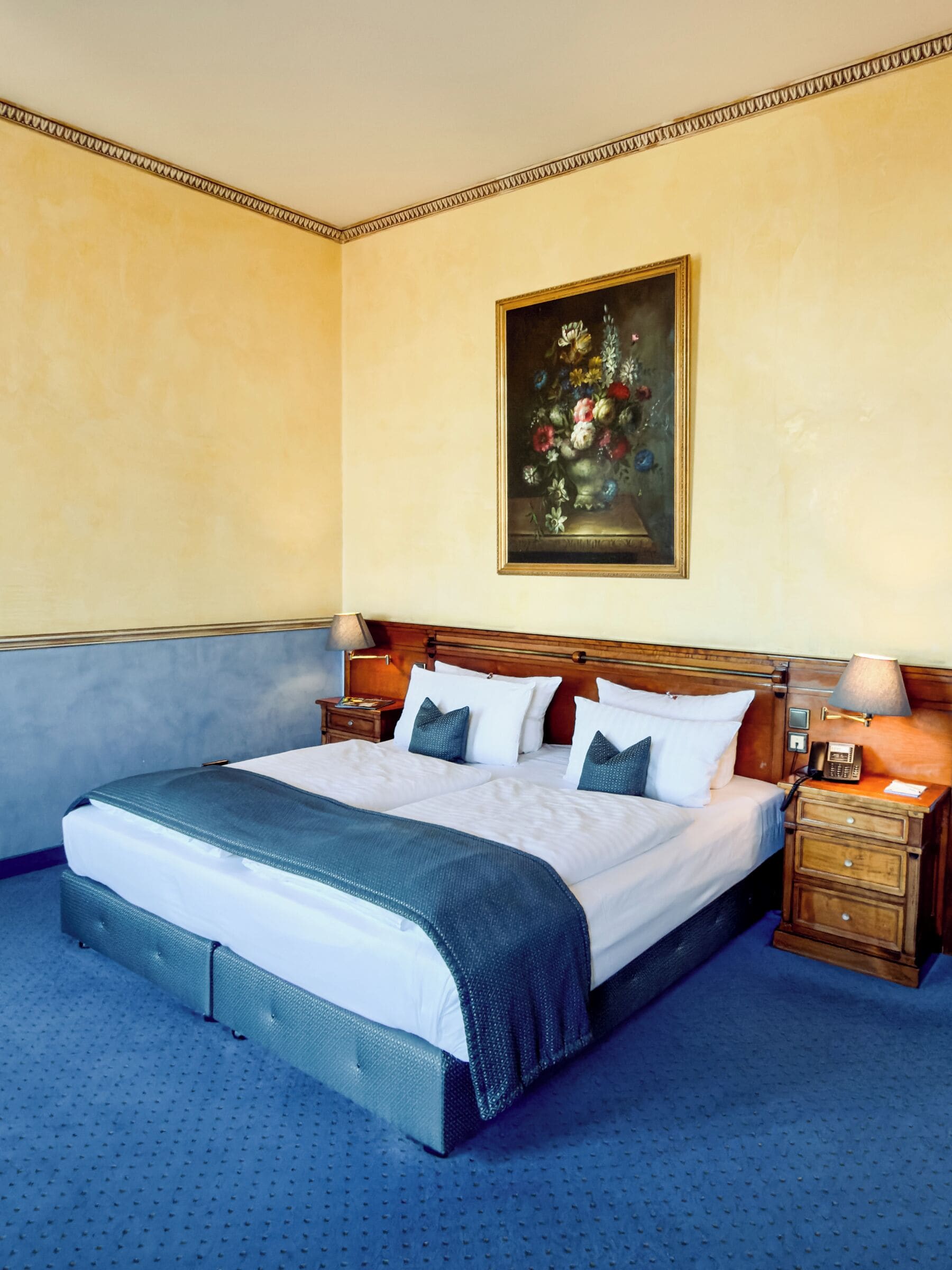 Una cama grande con cabecera de madera, dos mesitas de noche con lámparas, y un cuadro de flores en la pared.