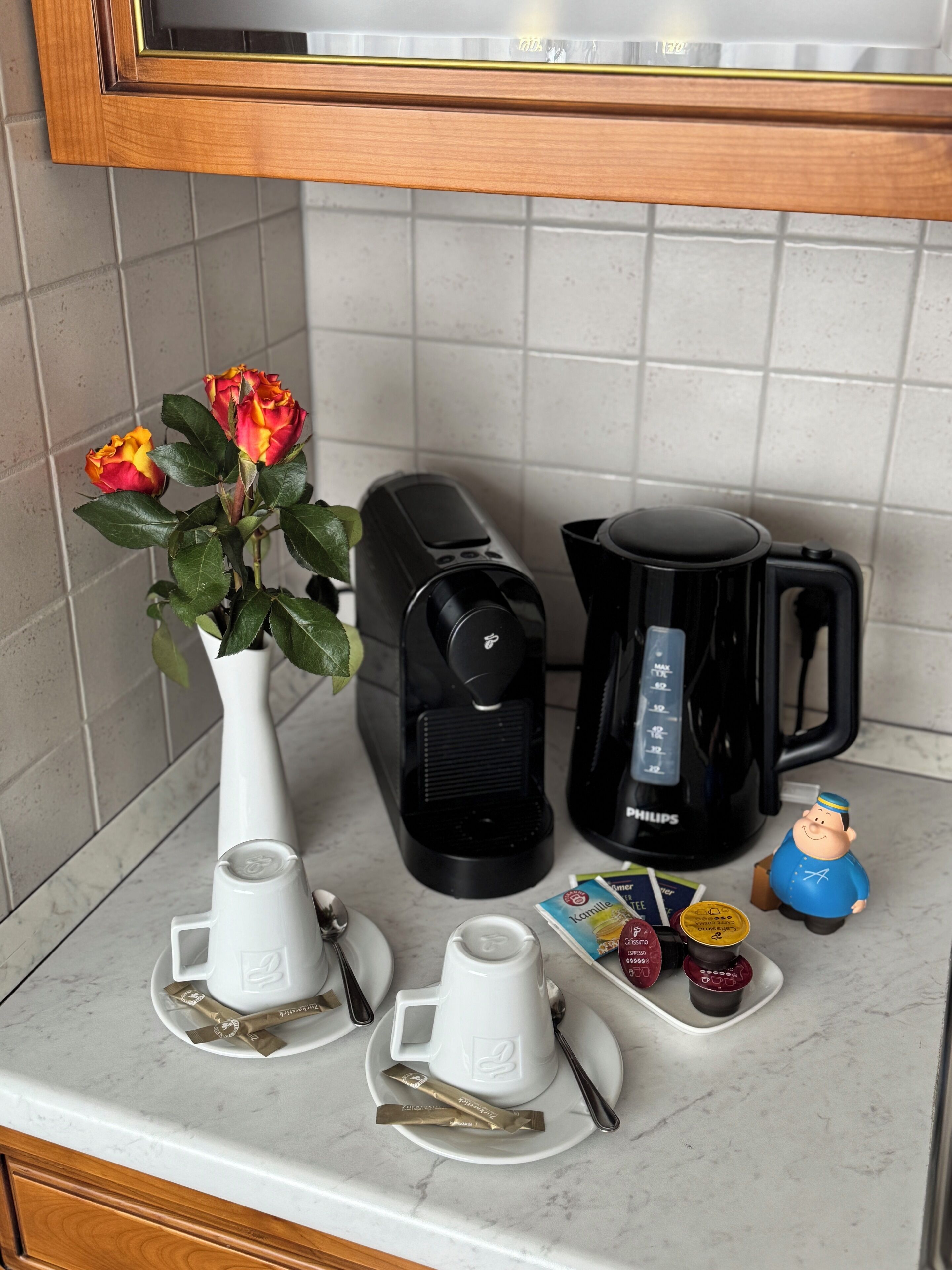 A kitchen counter with a coffee maker, a coffee pot, a small figurine, and a vase with flowers.