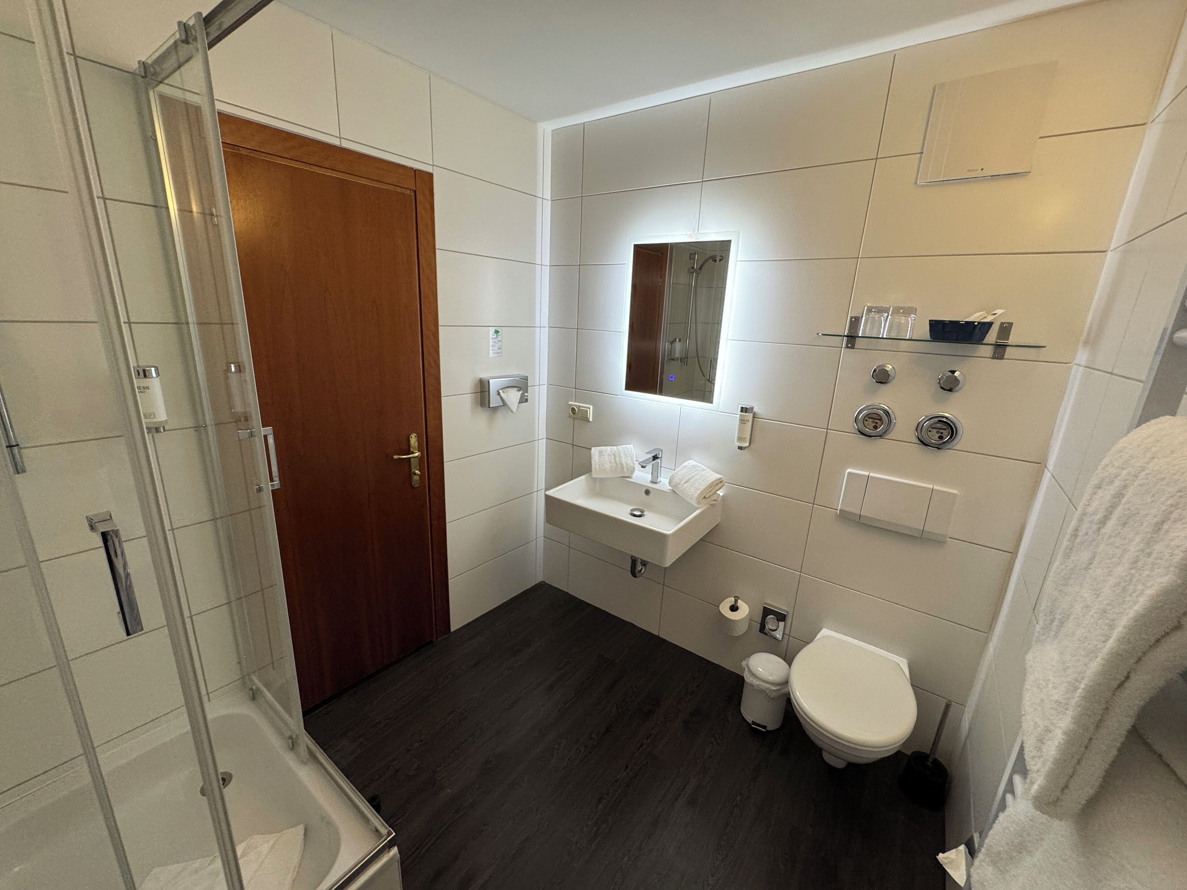 A modern bathroom with a glass shower enclosure, a wooden door, a white sink, a toilet, and a mirror.